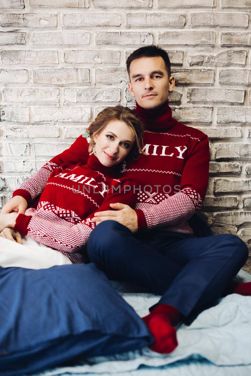 Portrait of young positive couple wearing in red sweaters with discription family. Happy boyfriend and girlfriend embracing and leaning on wall at studio. Wife and husband celebrating xmas.