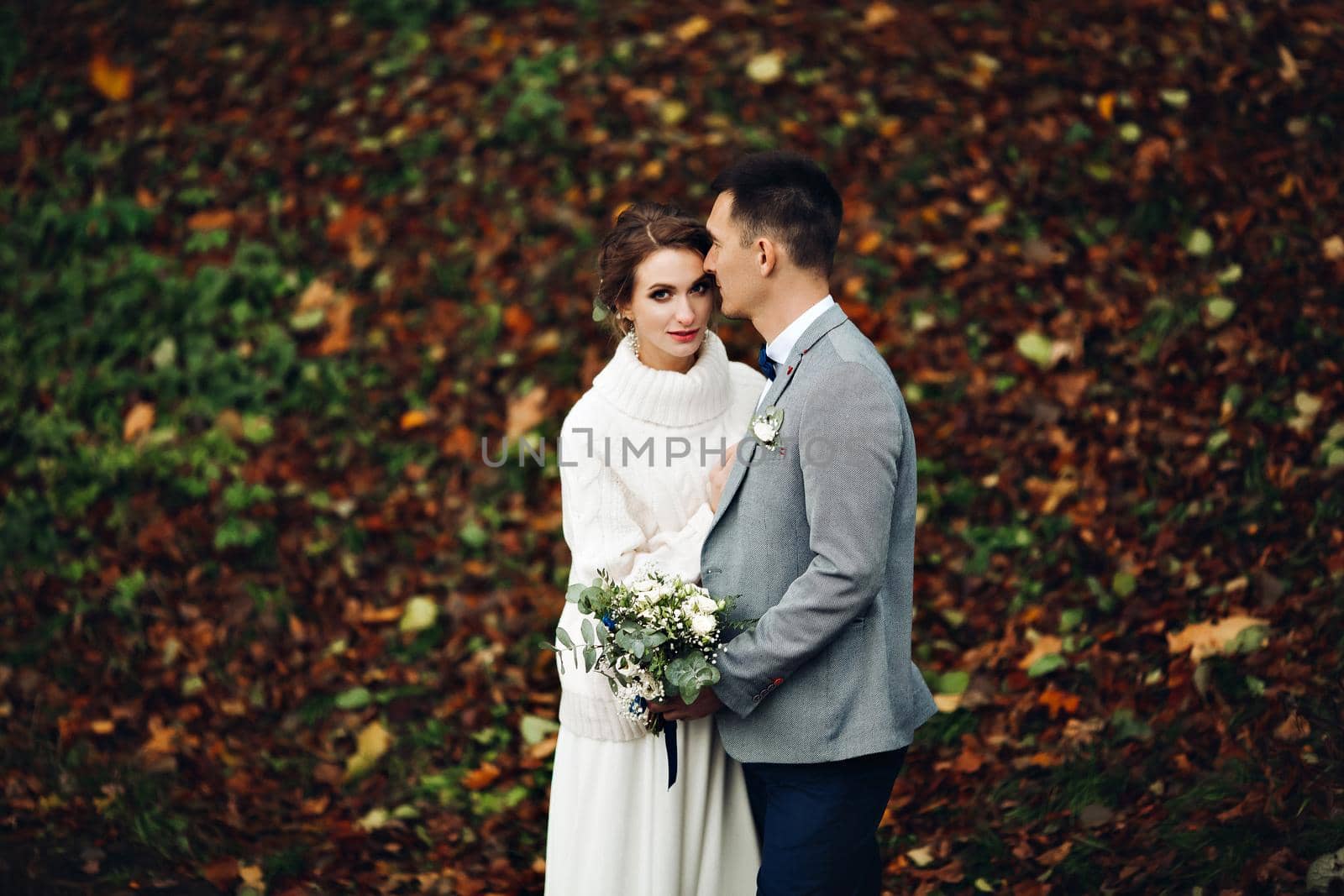 Portrait of groom and bride posing at autumn forest, among leaves, embracing and looking at camera. Gorgeous and beautiful bride and elegance groom touching by face. Conceptual wedding.