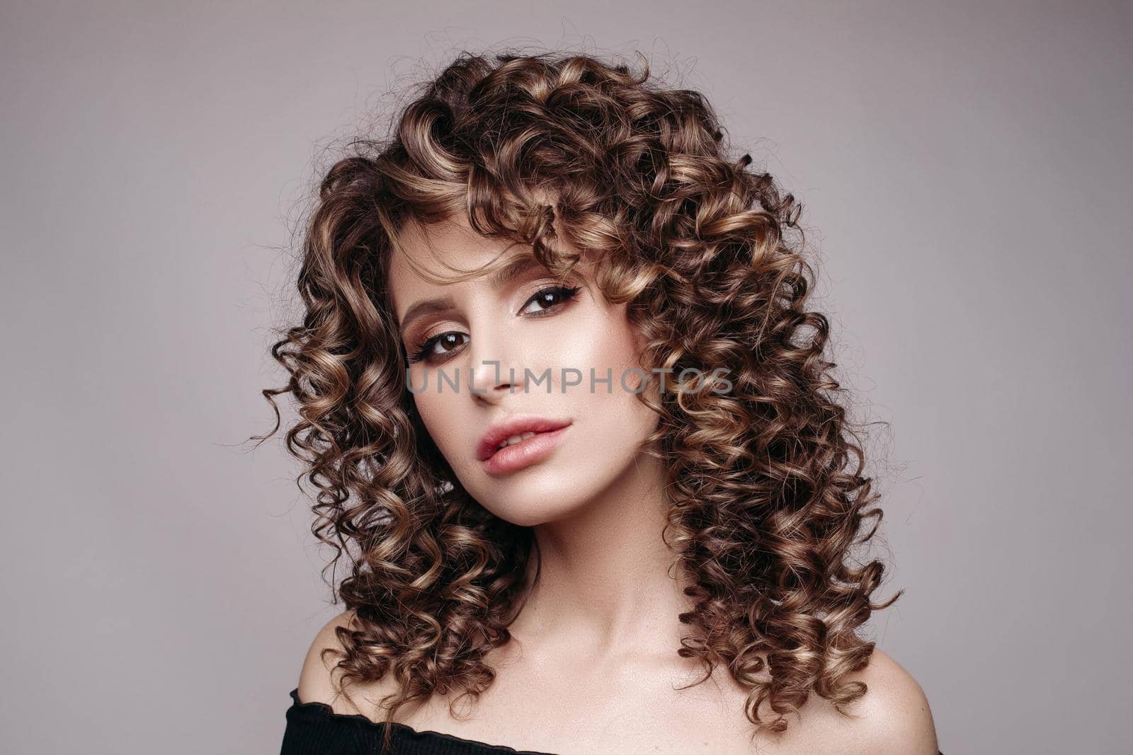 Studio close up of stunning young model with professional make-up and frizzy hair in black with open shoulders. She is looking at camera with passion. Isolate.