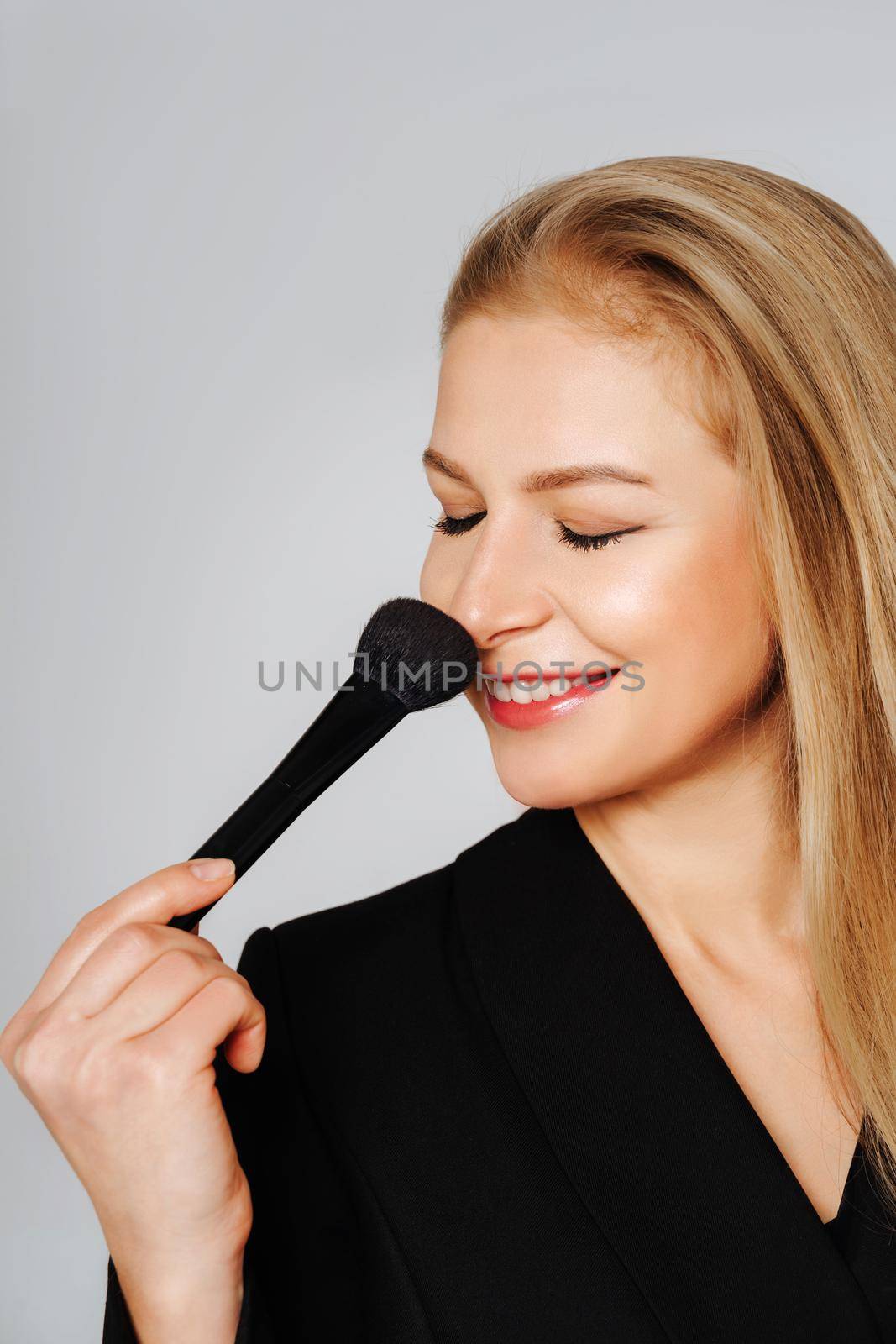 Beautiful middle aged woman waving one makeup brush, winking at the camera and smiling. Blond hair and a black jacket on a light background. by Matiunina