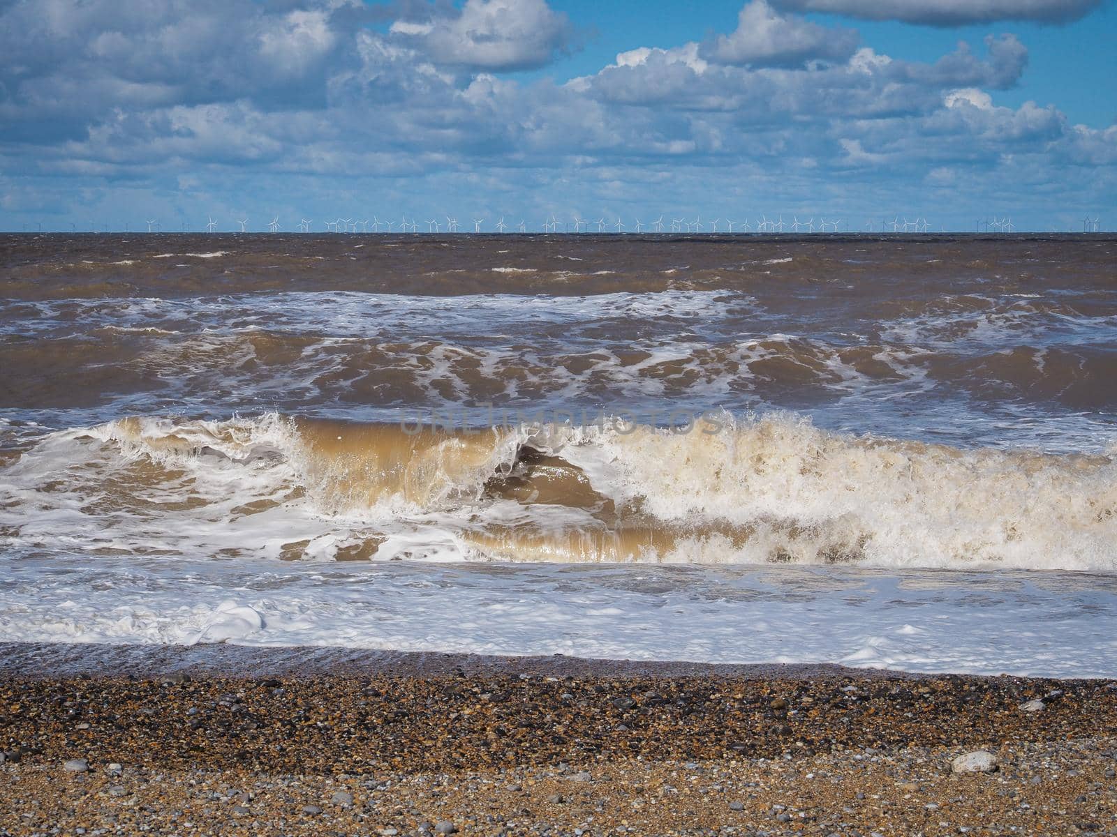 Rolling waves crash onto the beach with the white wind turbines of a massive wind farm on the horizon, Blakeney Point, Cley next the Sea, Norfolk, UK