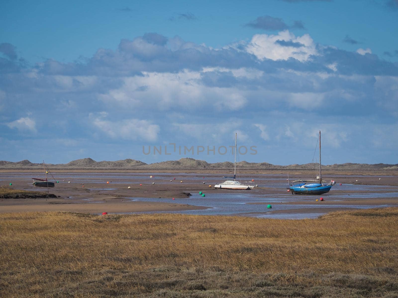 Boats aground waiting for the tide to come in on the expansive salt marshes under blue sky and white clouds, Blakeney National Nature Reserve, Norfolk, UK