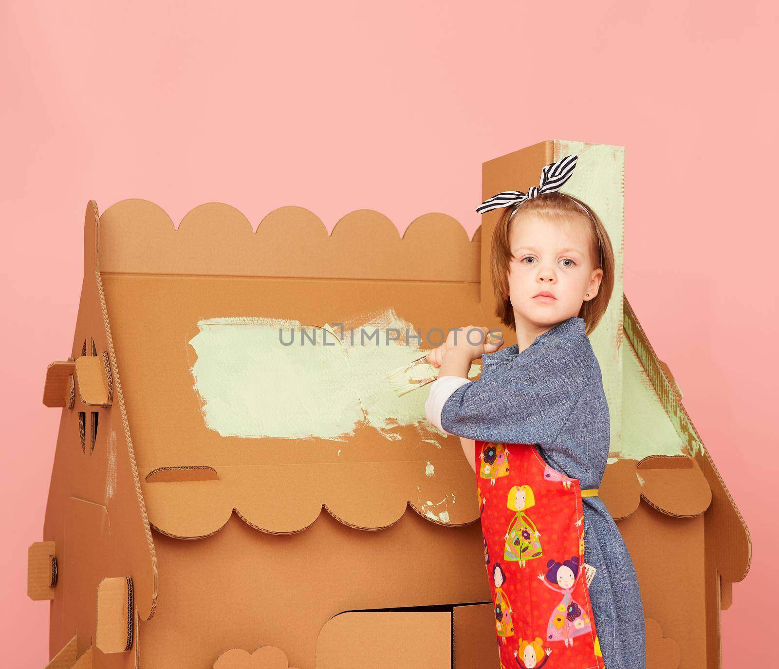 Young Girl Painting Big Cardboard House on over pink background.