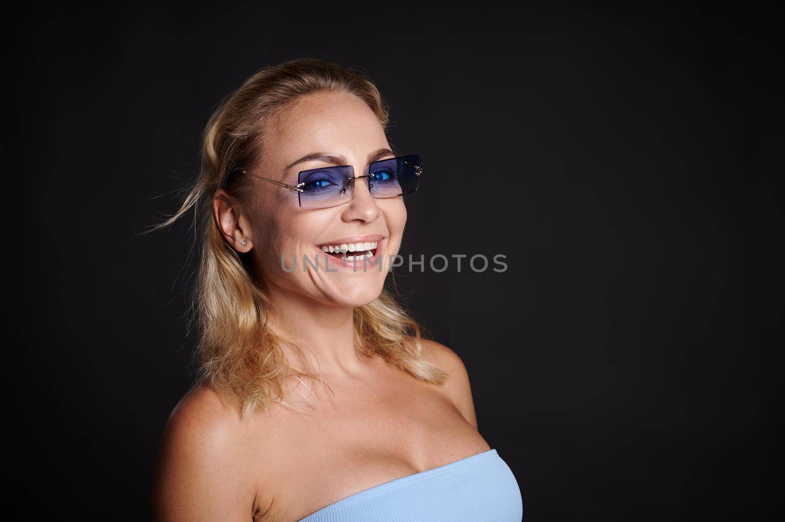 Beautiful European blonde woman in sunglasses smiles, looking at camera, isolated on black background.