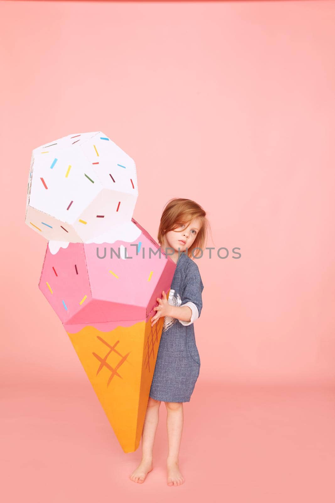 Portrait of a pretty little girl celebrating birthday and holding big ice cream toy in hands on over pink background.