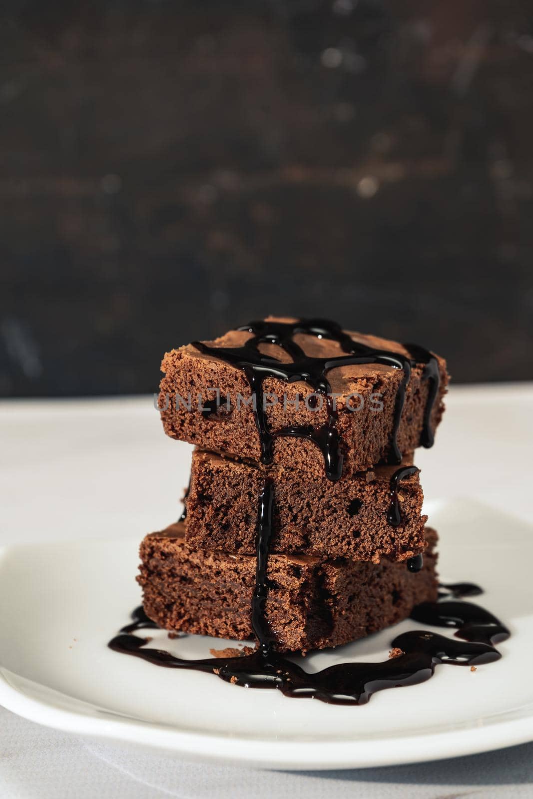 pile of chocolate brownies with strands of chocolate sauce on a white plate on a marble table. vertical orientation. Copy space.
