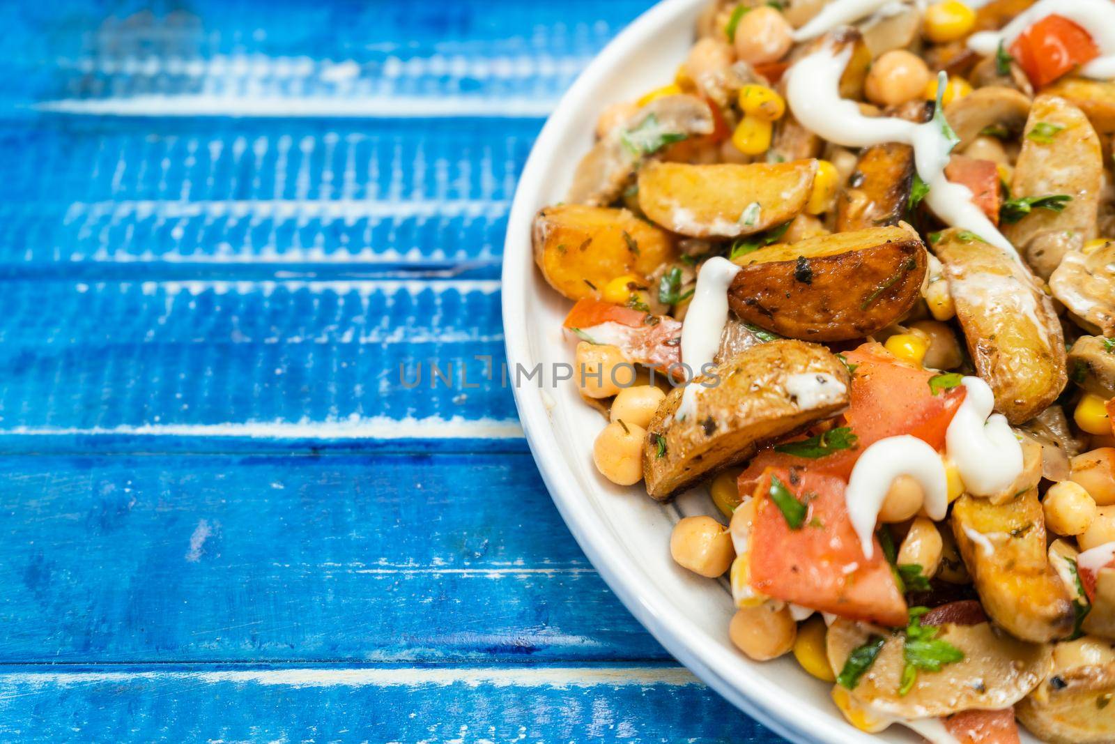 A delicious potato, chickpea, tomato and mushroom salad with parsley and aioli in an isolated plate on a blue rustic table. Healthy, homemade, vegan food. Partial view, copy space