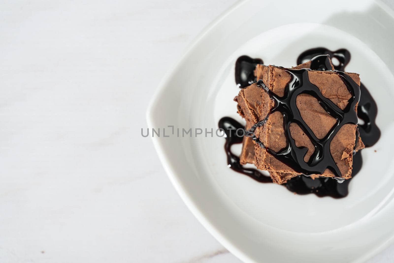 pile of chocolate brownies with strands of chocolate sauce on a white plate on a marble table. Top view. Copy space.