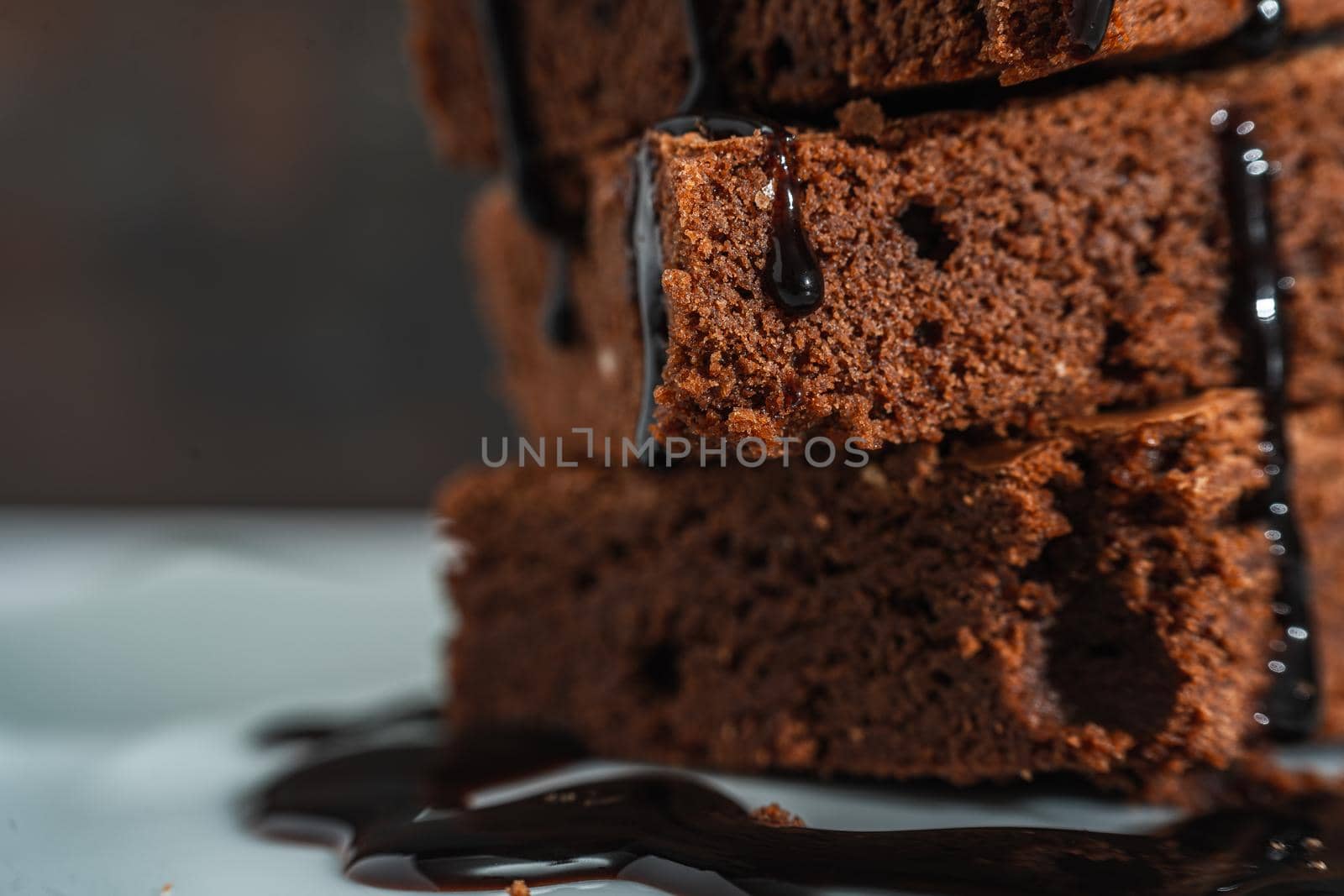 Very close shot of a Pile of Chocolate Brownies with chocolate strands falling on a white plate and dark background. Normal view.