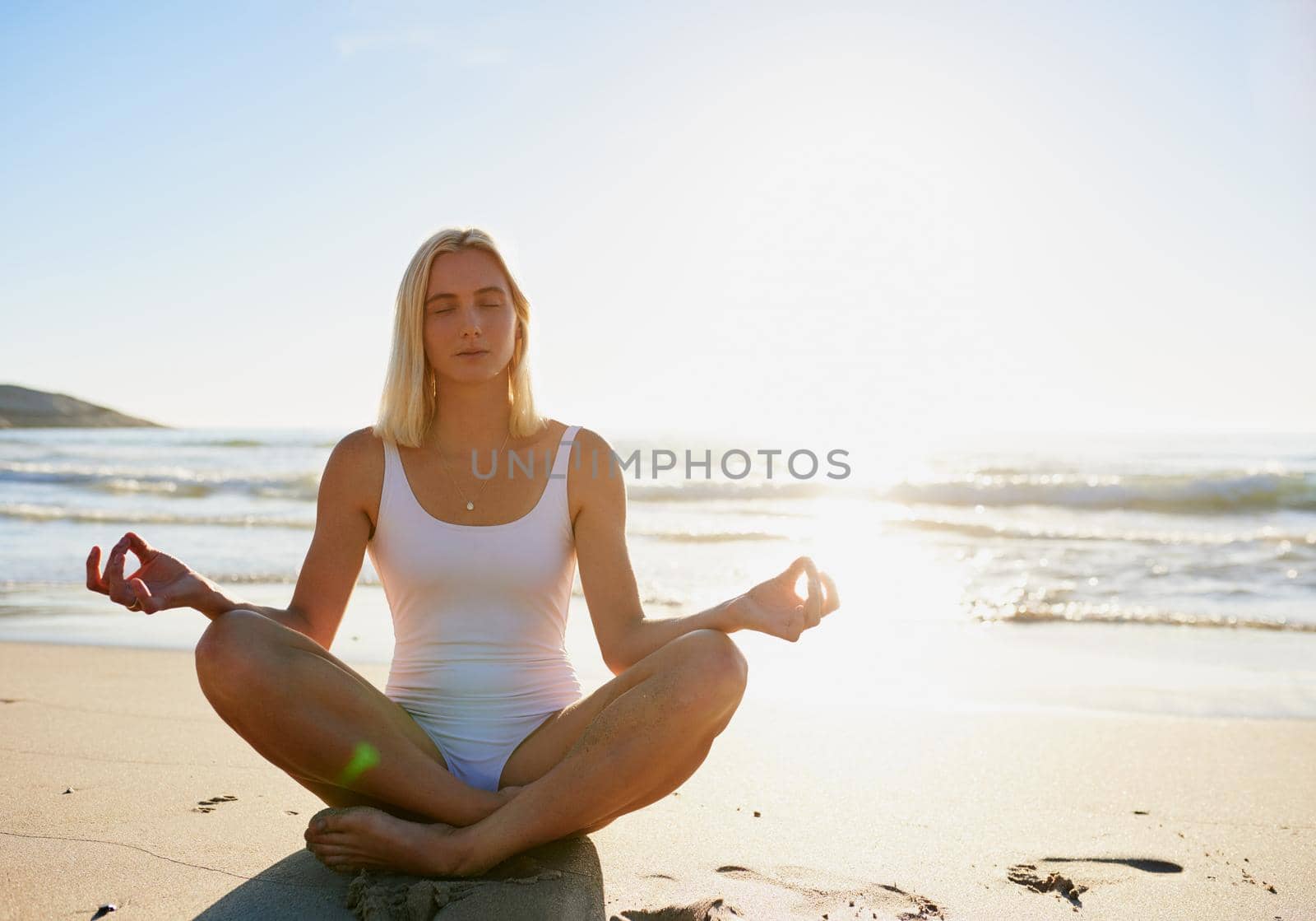 Full length shot of an attractive young woman sitting with her legs crossed and meditating on the beach.