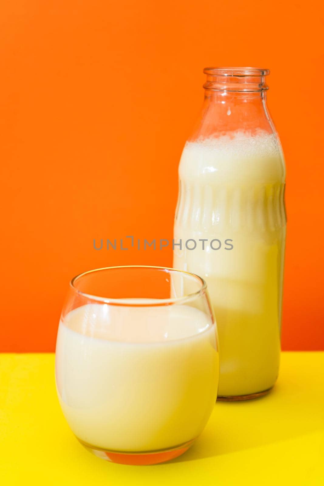 Glass bottle and large glass with milk on a yellow table with orange background. Copy space. vertical orientation