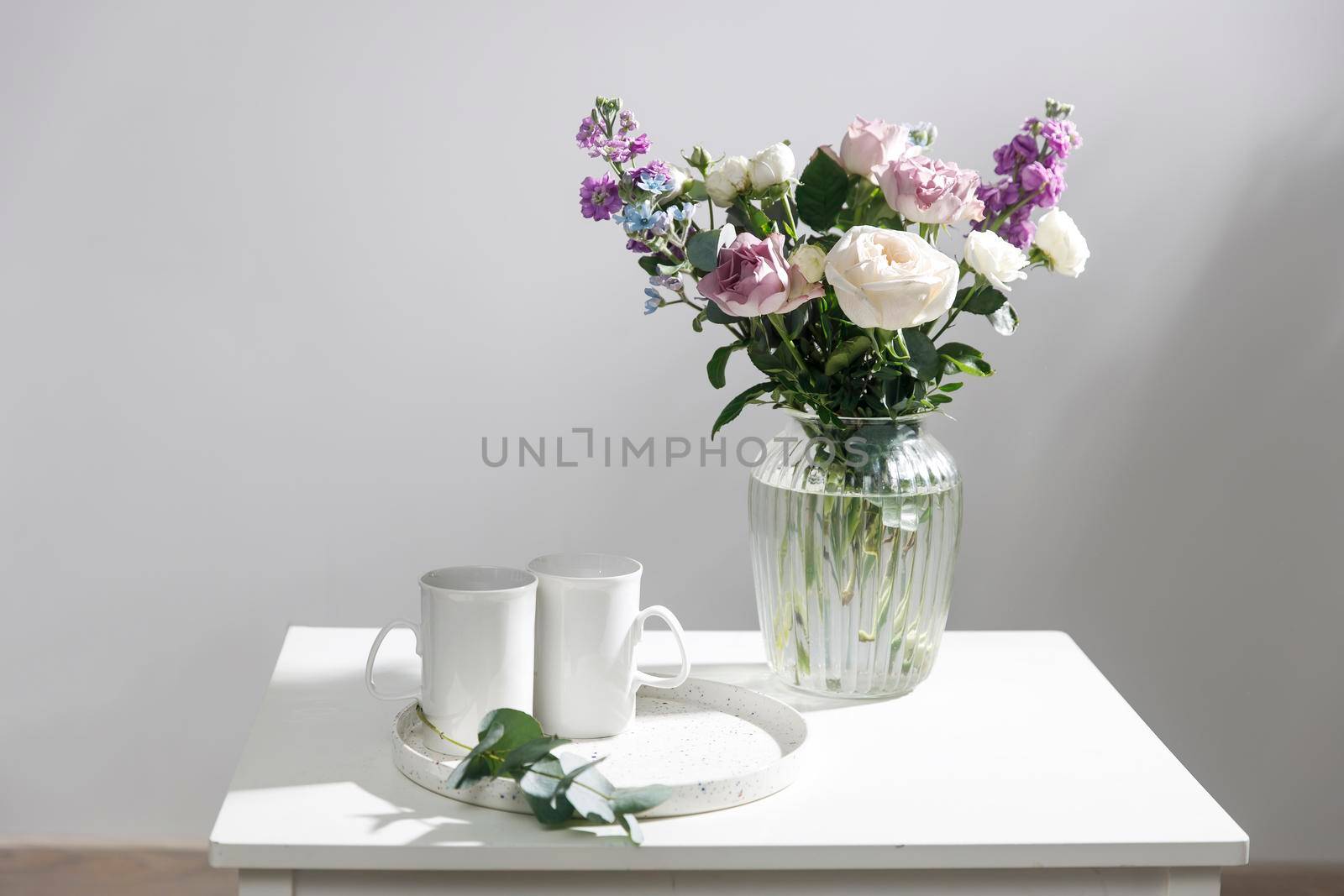 Bouquet of hackelia velutina, purple and white roses, small tea roses, matthiola incana and blue iris in glass vase is on white coffee table with two tall cups for tea. Grey wall by elenarostunova