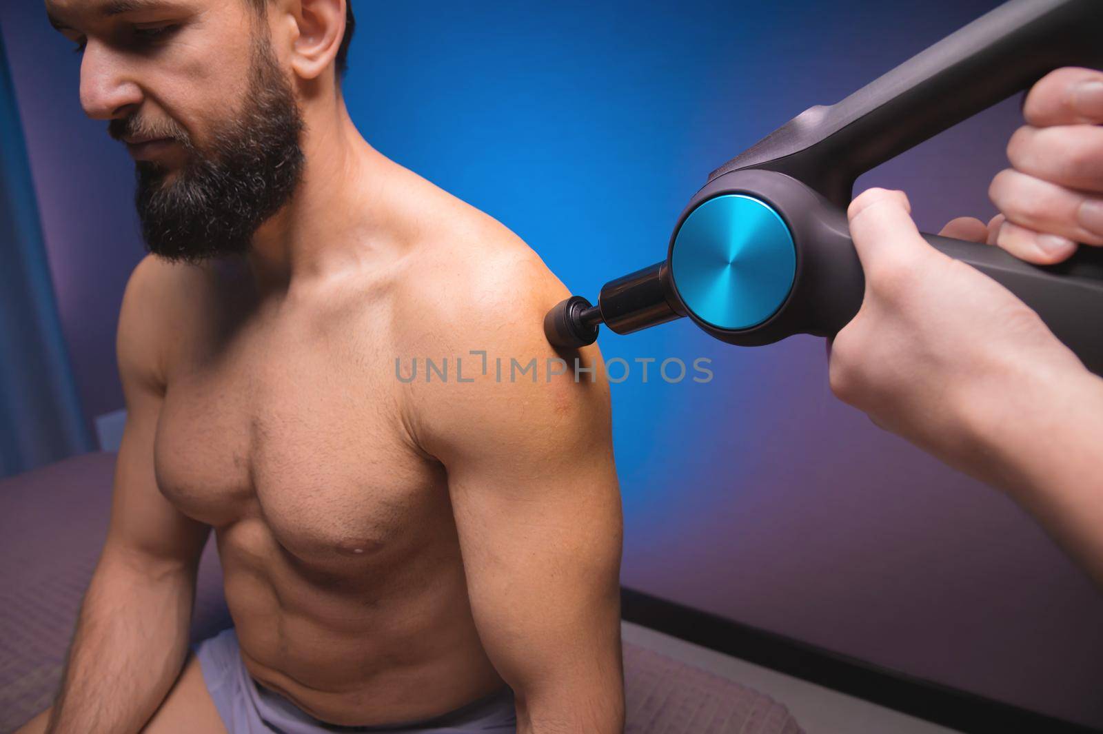 Bearded caucasian man getting percussion shoulder massage by a massager in a professional massage parlour. shock wave muscle recovery technique.