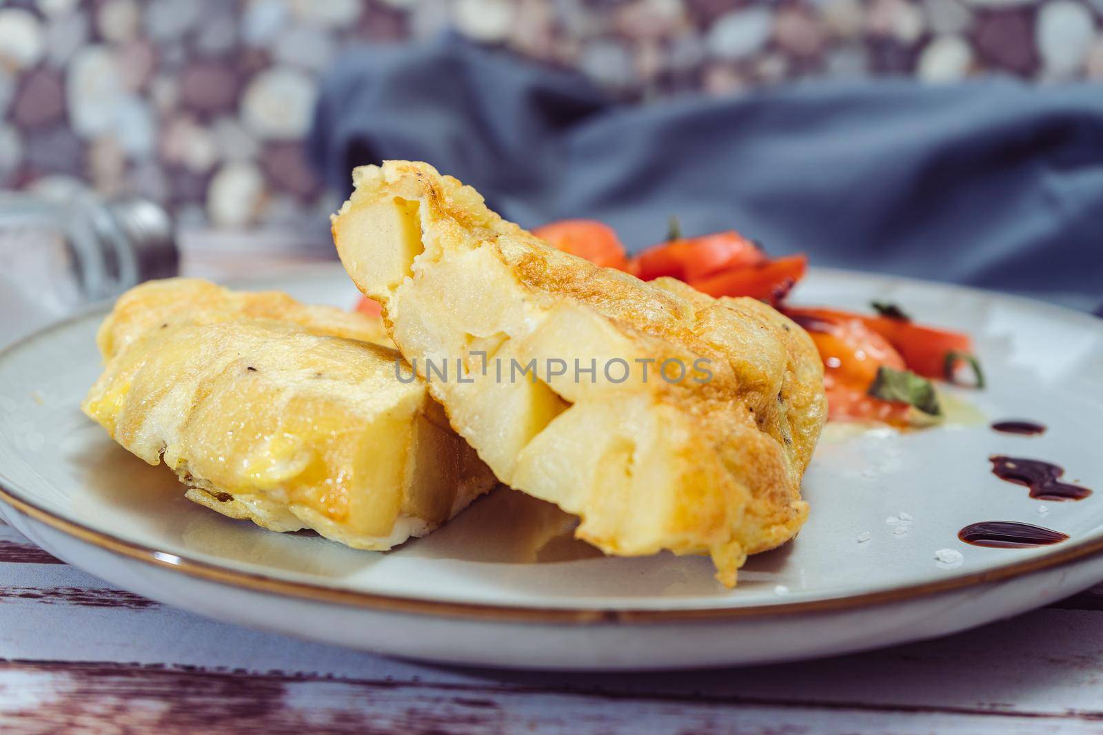 Normal view of a plate with two portions of Spanish potato omelette and sliced tomatoes with olive and sherry vinegar on a wooden rustic table.