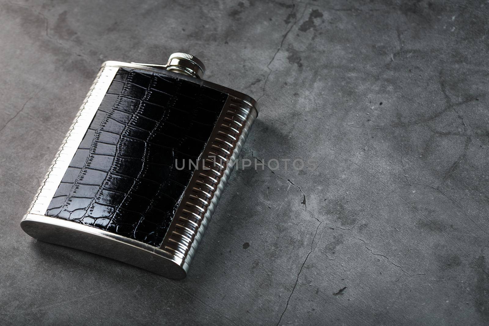 Pocket flask made of steel in leather finish with an alcoholic drink on a dark background with copy space