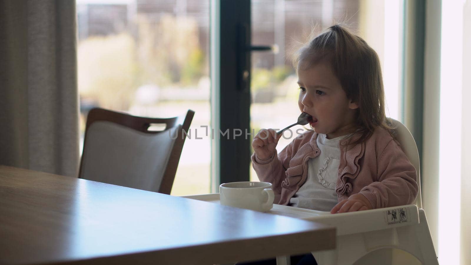 Closeup of young baby in white feeding high chair, kid is trying to eat himself, happy child with food stained face, little Girl eats porridge with spoon. High quality feeding up breakfast in kitchen.