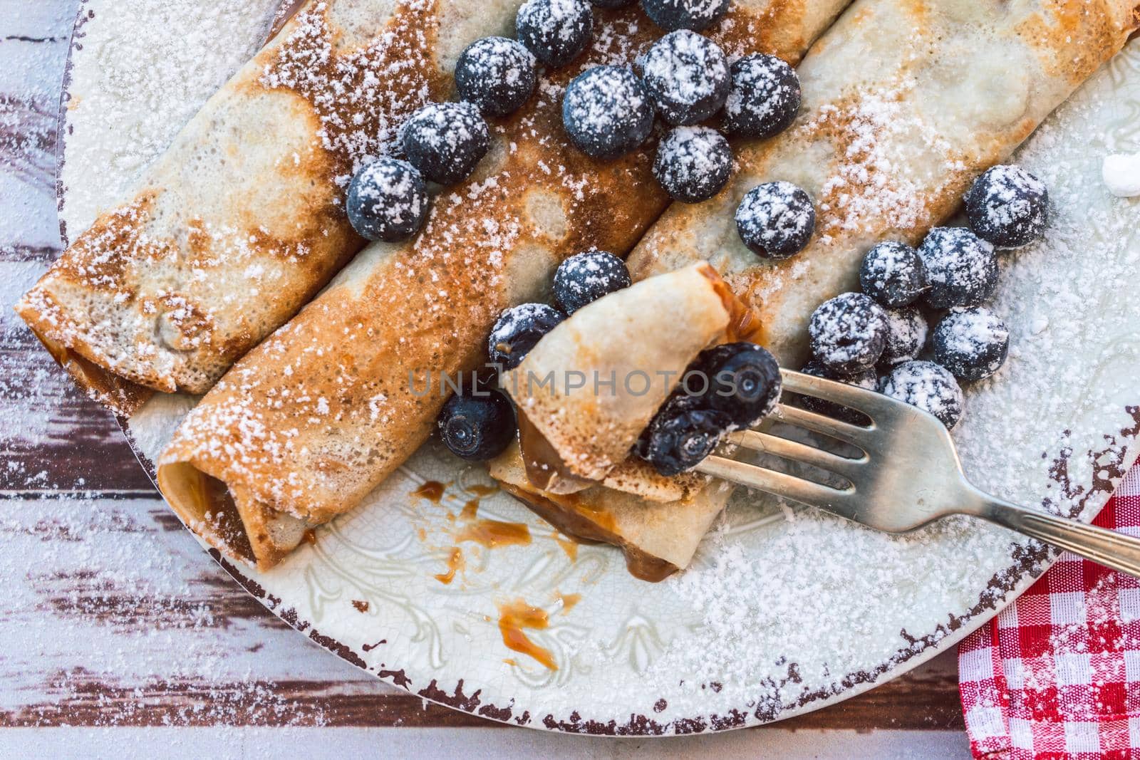 Dish with delicious homemade pancakes or crepes filled with dulce de leche. Fork with a bite and some spiked blueberries. Close up. high view.