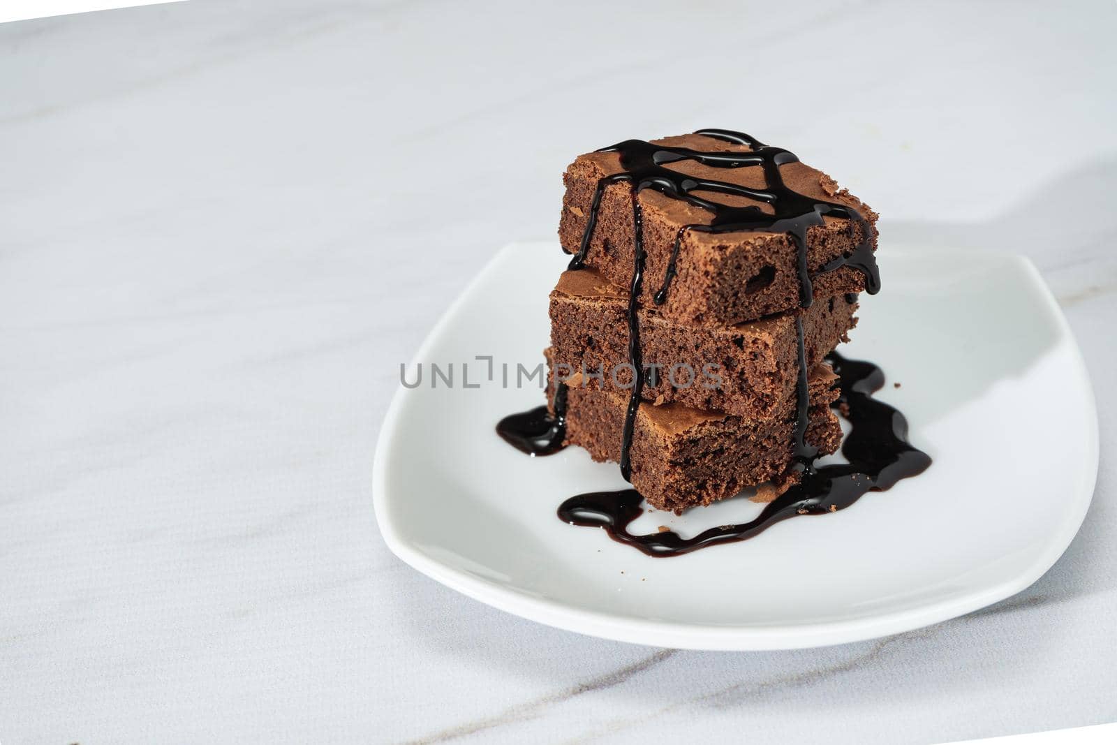 pile of chocolate brownies with strands of chocolate sauce on a white plate on a marble table. Copy space.