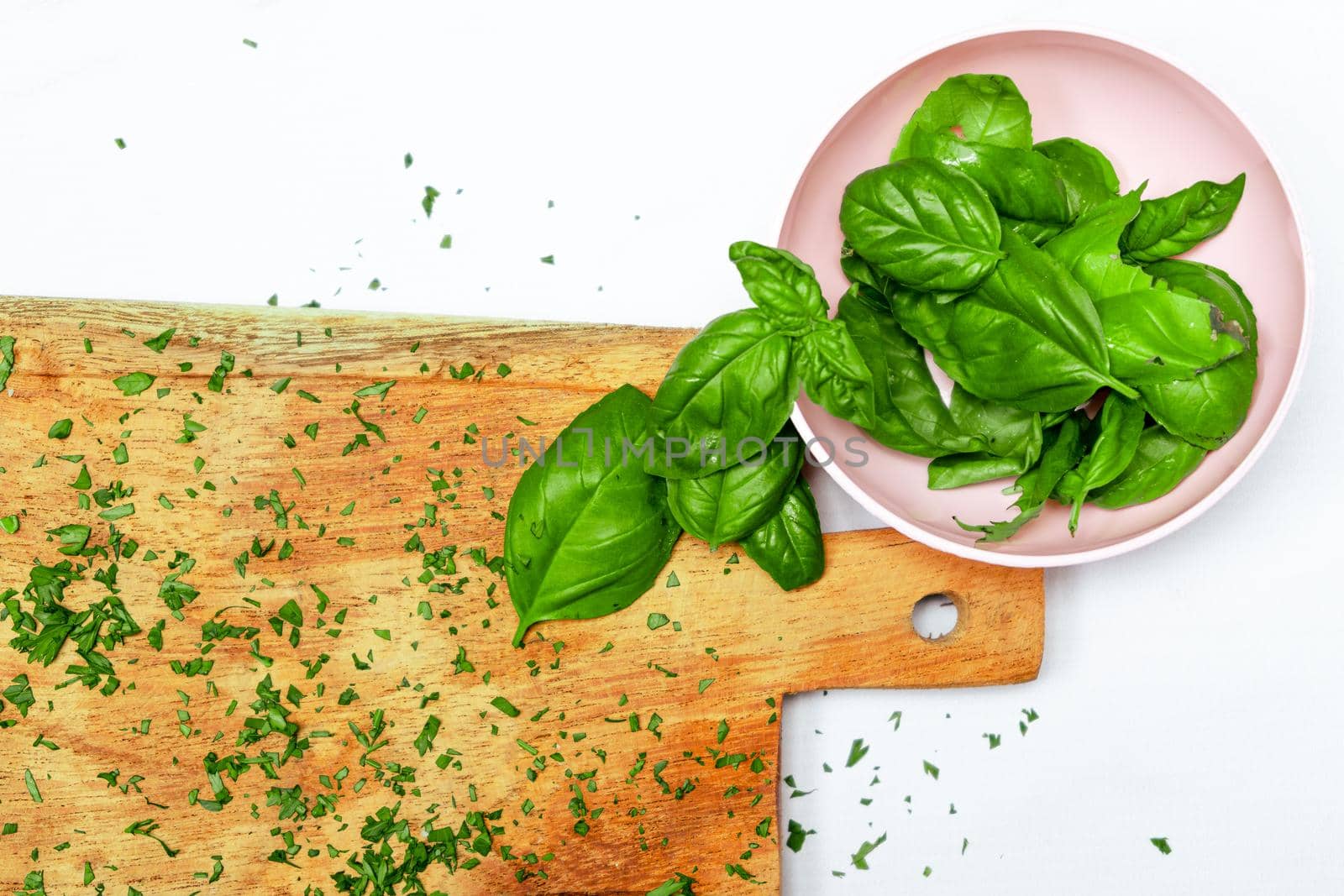 Fresh basil leaves in a small bowl next to a wooden cutting board on a white surface. Fresh, healthy and natural food concept. Copy space.