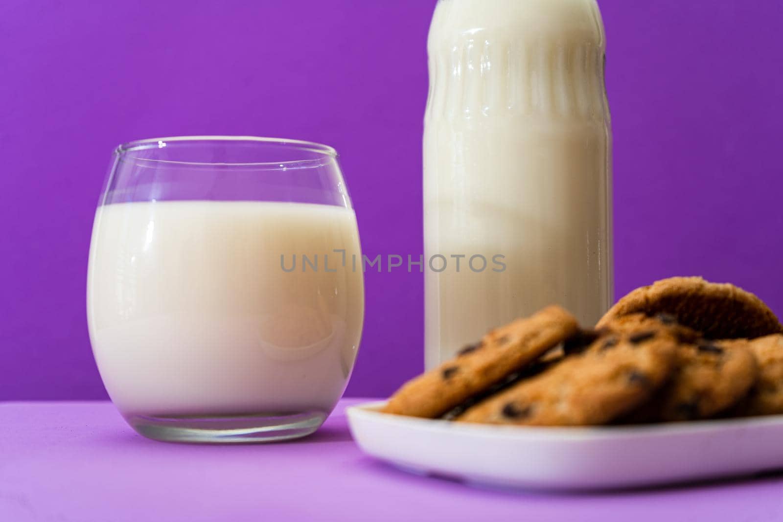 Close-up of a Glass bottle and large glass with milk and some sweet chocolate chip cookies in a purple or violet environment. Copy space.