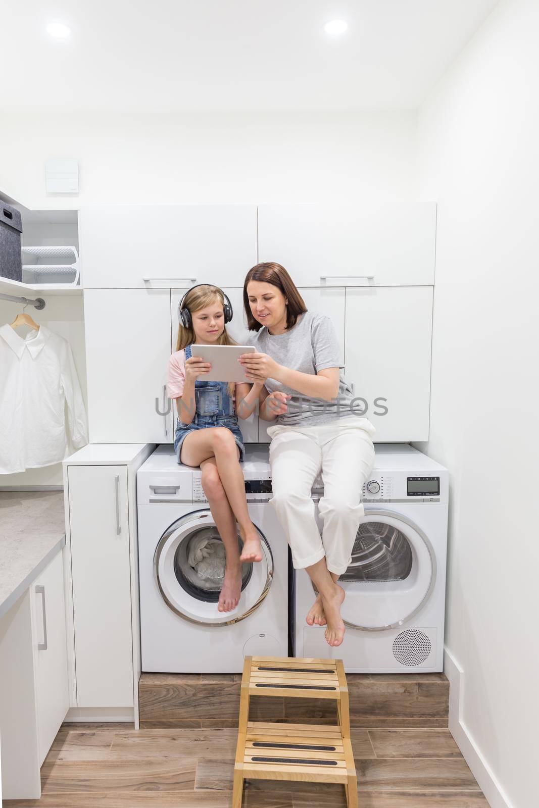 Young mother and her daughter are playing with tablet in laundry room sitting on washing machine by Mariakray