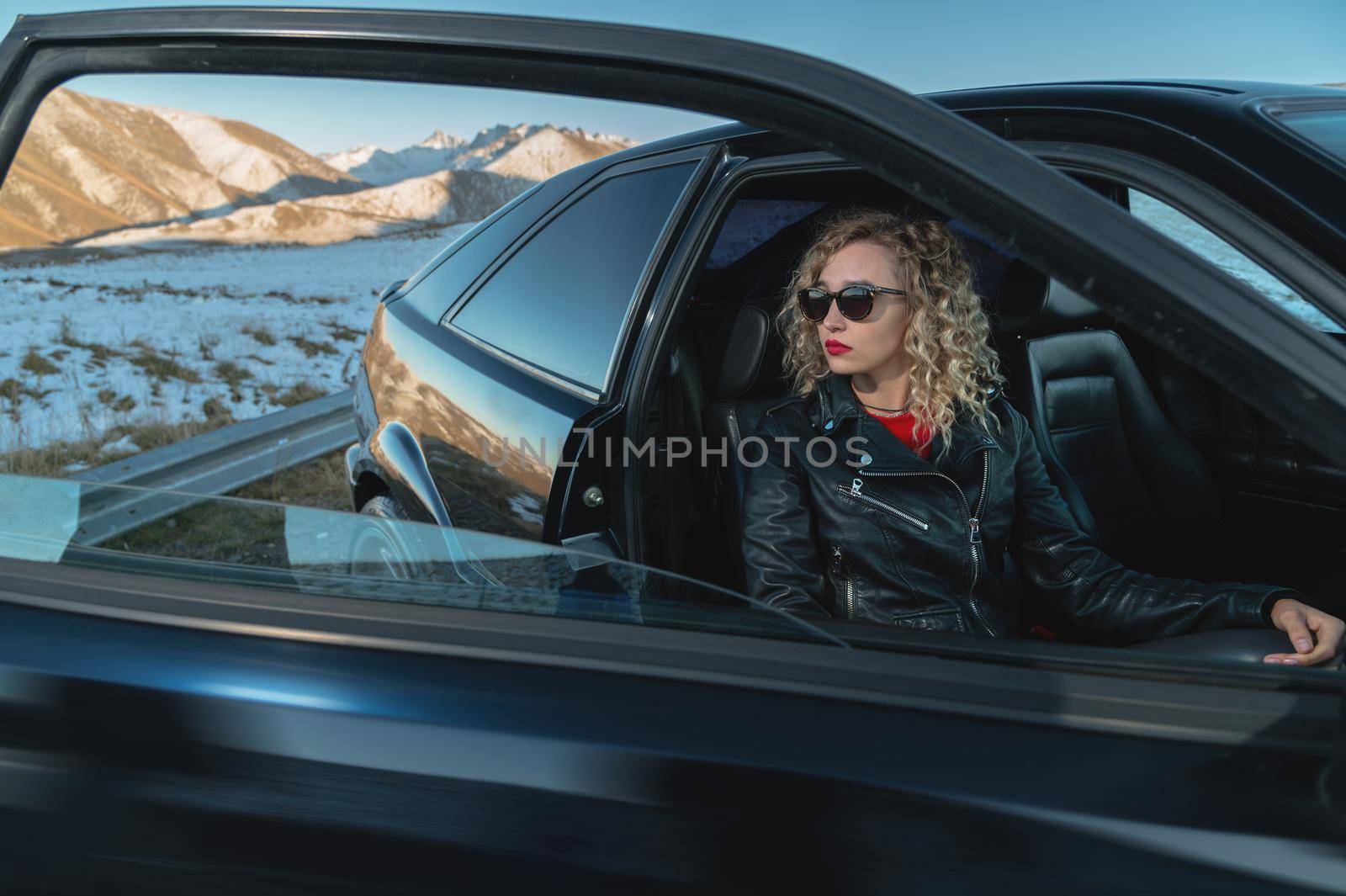 Portrait of a stylish attractive Caucasian young woman getting out of a retro sports car in the mountains at sunset. Travel concept copy space.