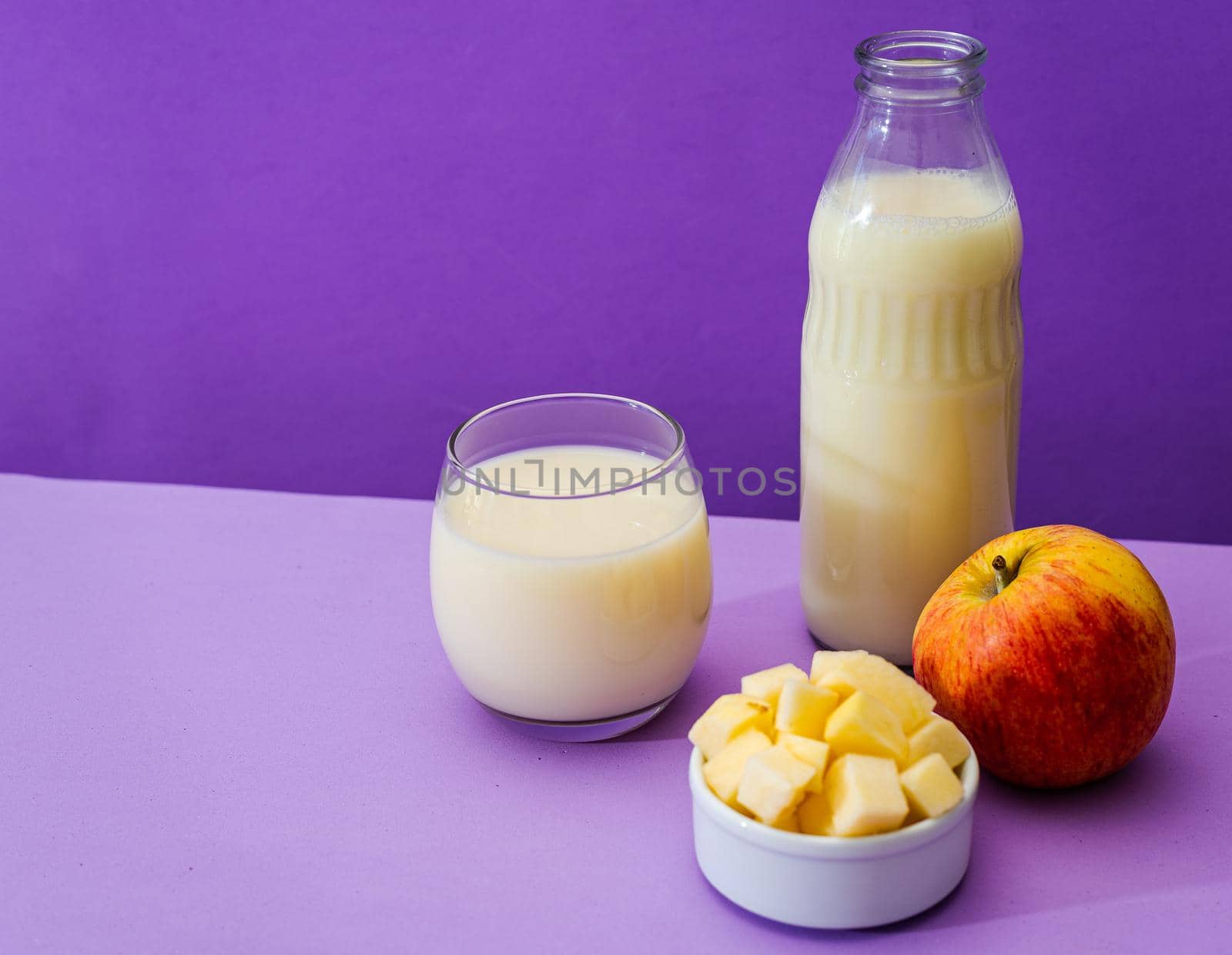 Glass bottle and large glass with milk and a whole red apple next to a bowl with apple cubes in a purple or violet environment. Copy space. Landscape orientation.
