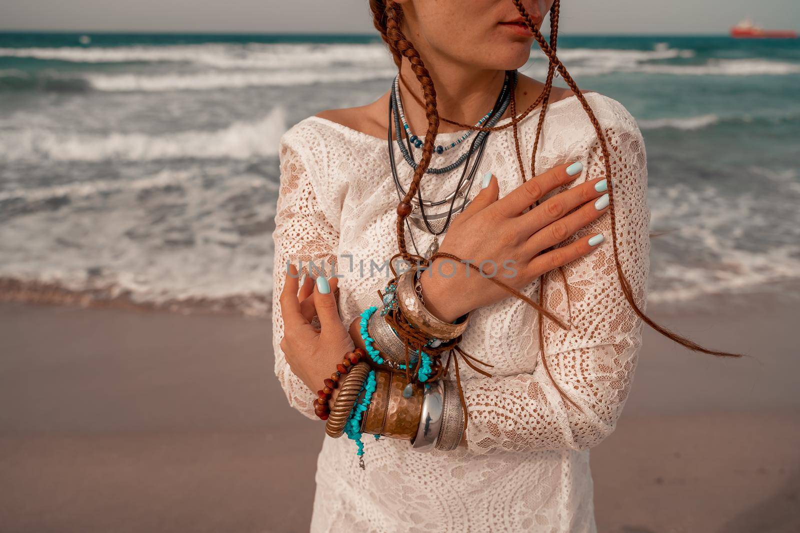 Model in boho style in a white long dress and silver jewelry on the beach. Her hair is braided, and there are many bracelets on her arms. by Matiunina