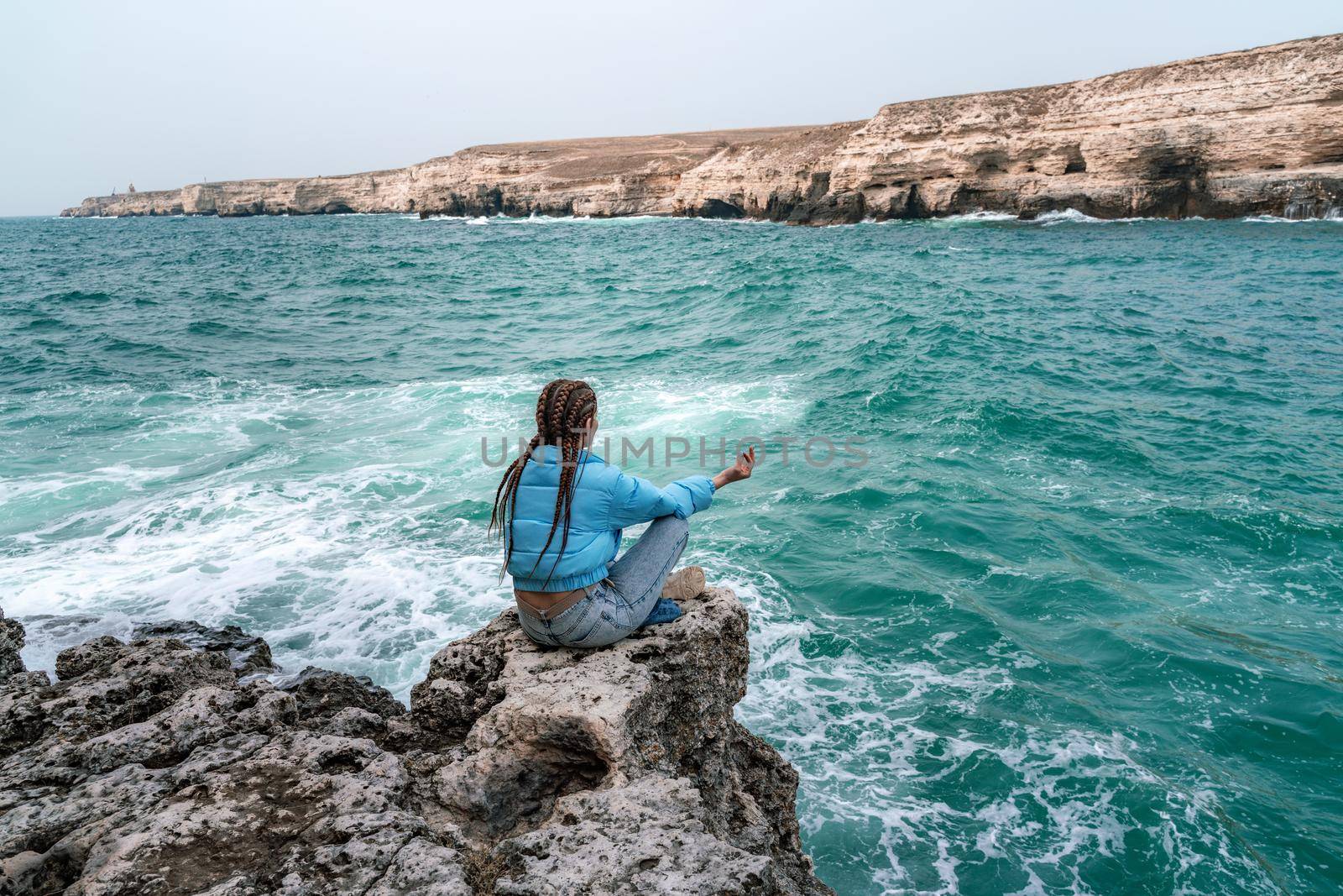 A woman in a blue jacket sits on a rock above a cliff above the sea, looking at the stormy ocean. Girl traveler rests, thinks, dreams, enjoys nature. Peace and calm landscape, windy weather. by Matiunina