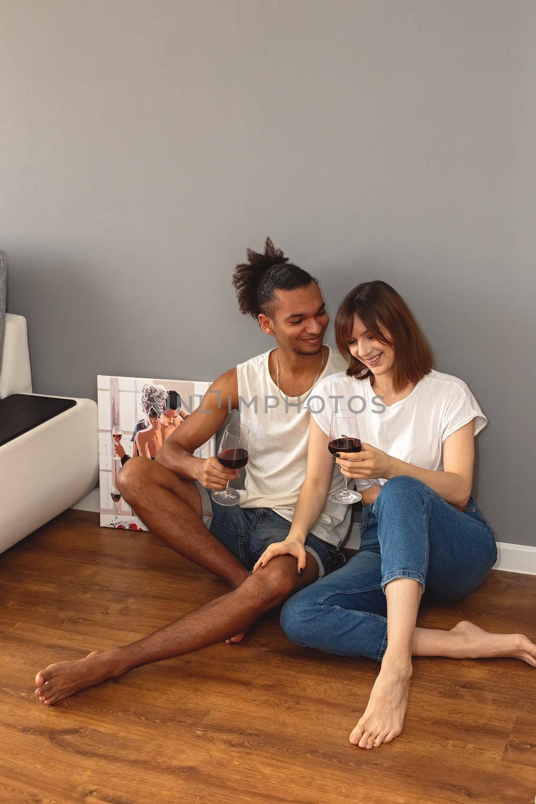 Stylish, modern, multiracial couple, a guy and a girl in white t-shirts and blue jeans, sit at home on the floor against a gray wall, hold glasses of red wine in their hands, happy. copy space. Vertical