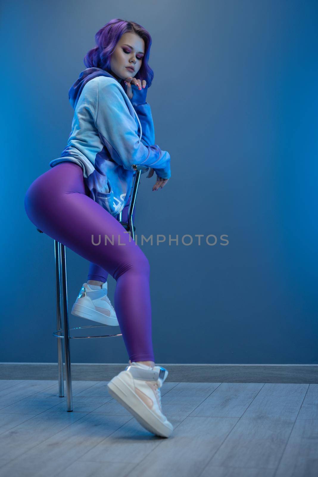 girl in stylish purple sportswear and with purple hair poses sexually on a bar stool