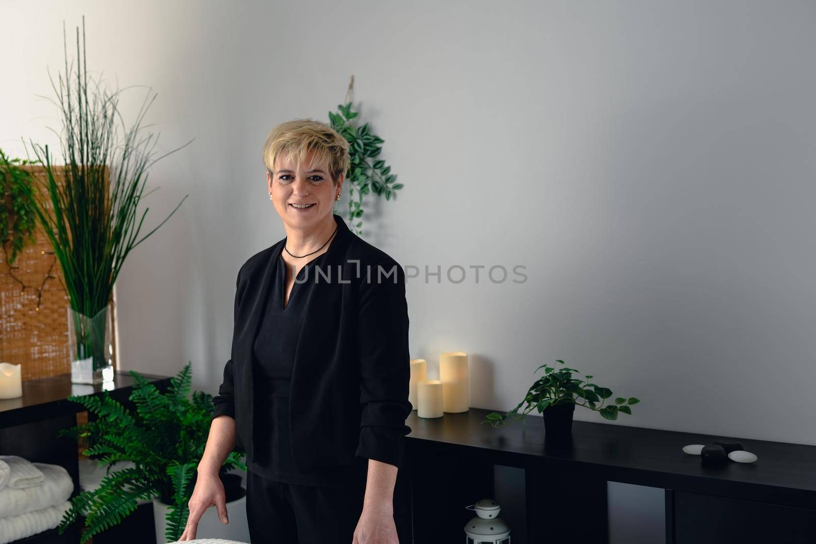 Portrait of a mature, smiling female beautician, dressed in black company uniform, posing standing in her small spa business, to start the day. Relaxing atmosphere and subdued lighting, decoration of plants and candles, background massage table and towels. Horizontal