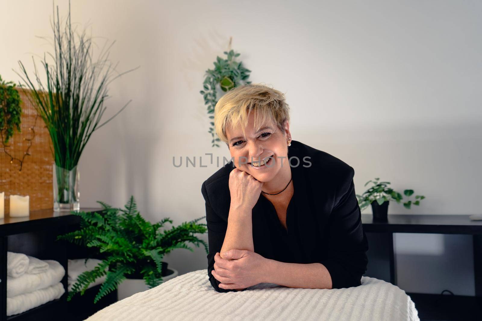 Portrait of a mature, smiling female beautician, dressed in black company uniform, posing standing in her small spa business, to start the day. Relaxing atmosphere and subdued lighting, decoration of plants and candles, background massage table and towels. Horizontal