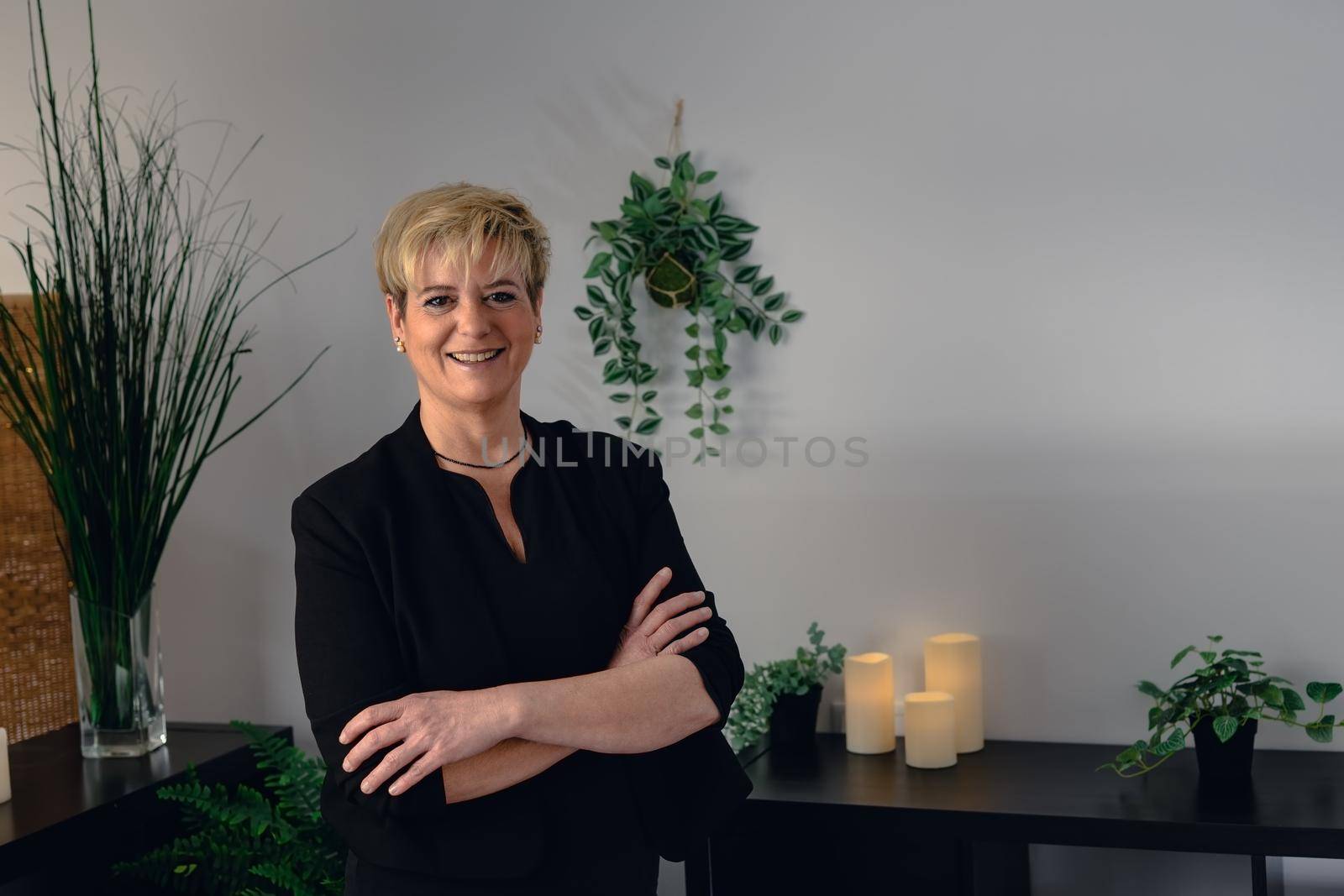 Portrait of a mature, smiling female beautician, dressed in black company uniform, posing standing in her small spa business, to start the day. Relaxing atmosphere and subdued lighting, decoration of plants and candles, background massage table and towels. Horizontal