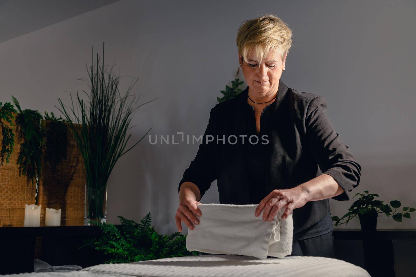 Portrait of a blonde-haired woman, beautician, mature, concentrated and professional, dressed in the black uniform of the company, preparing the massage table to start the day. Relaxing atmosphere and subdued lighting, decoration of plants and candles, background massage table and towels. Horizontal.