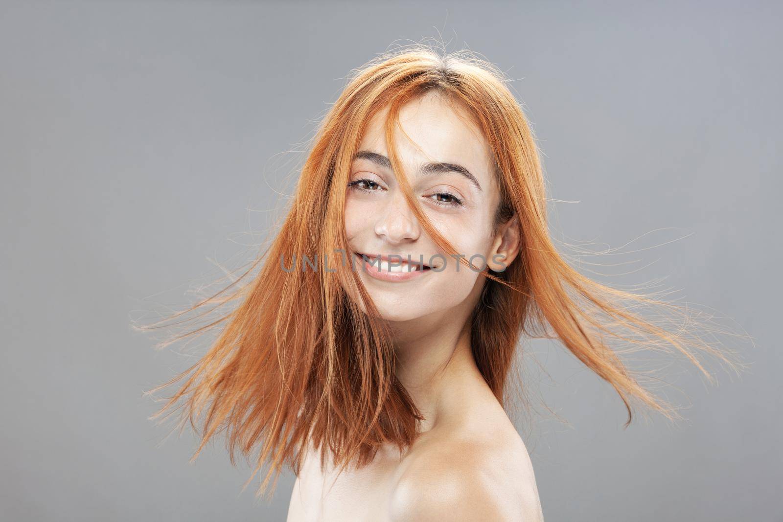 Beautiful dark burnt orange windy hair girl smiling. Studio portrait with happy face expression against gray background... by kokimk