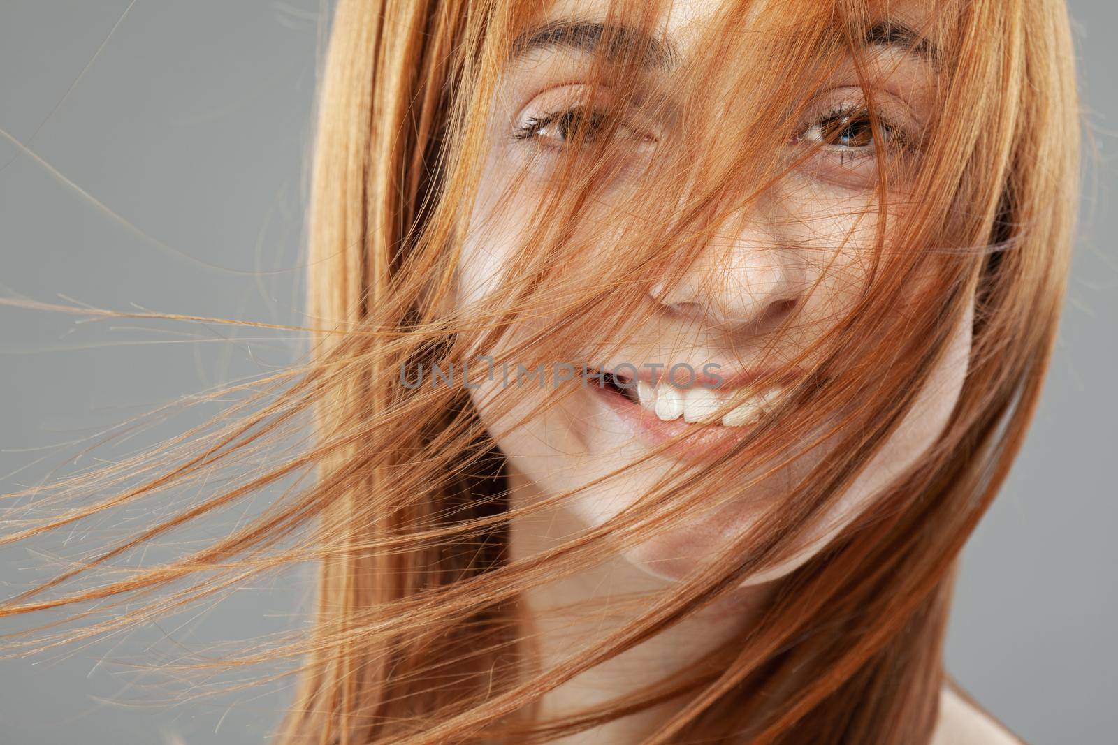 Beautiful dark burnt orange windy hair girl smiling. Studio portrait with happy face expression against gray background.