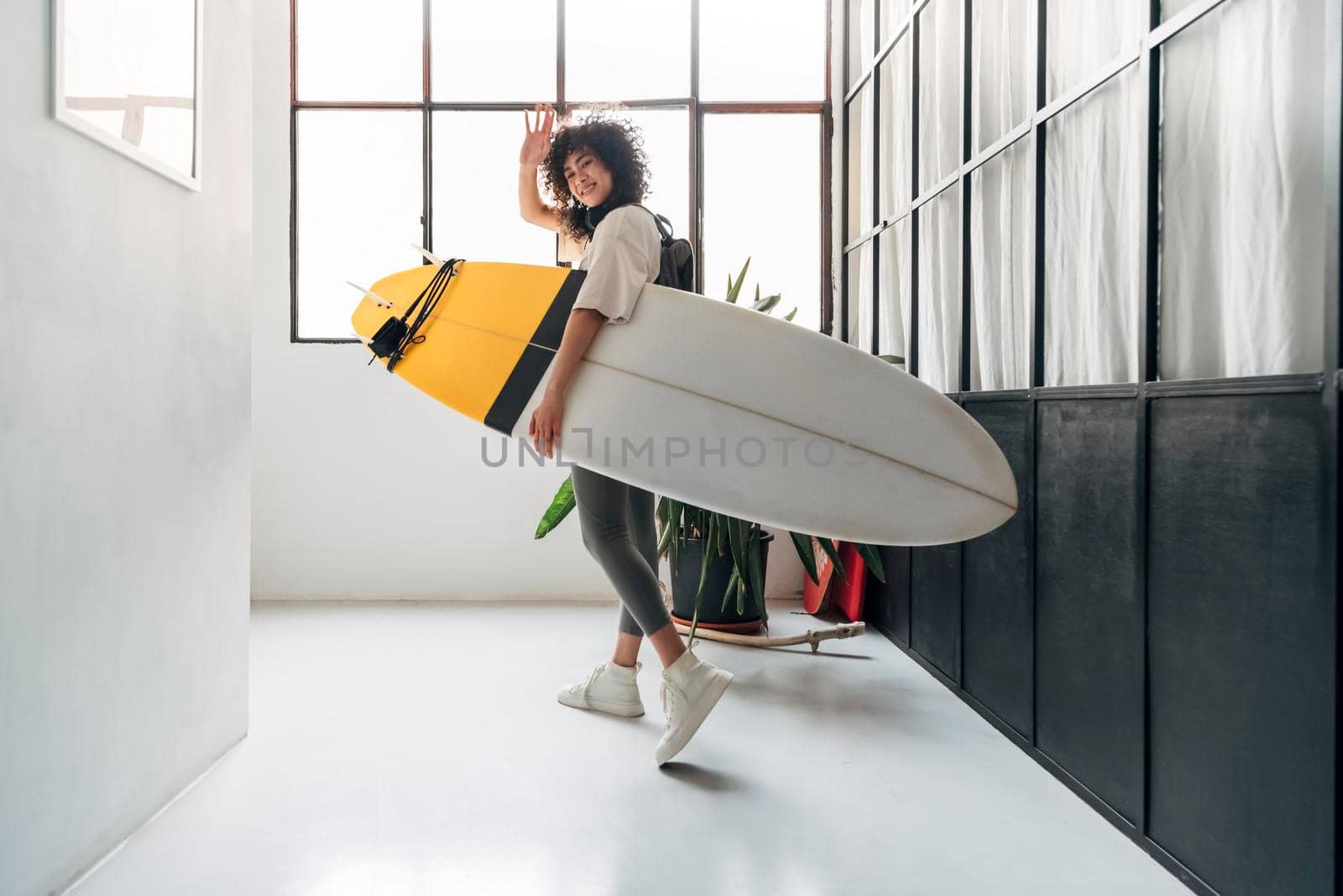 Young multiracial woman leaves apartment carrying surfboard to go surfing in the beach. by Hoverstock