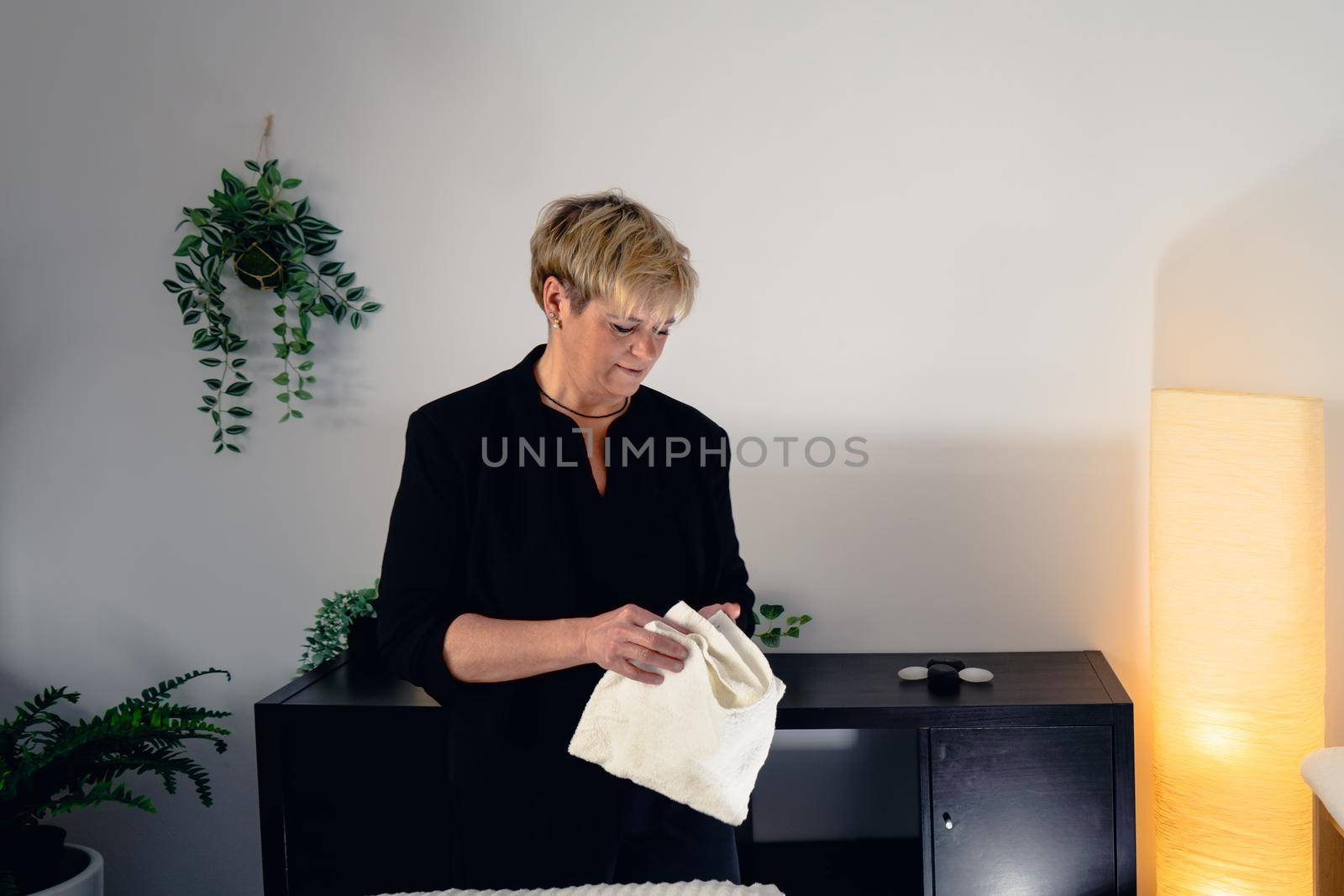 Portrait of a mature, smiling female beautician, dressed in black company uniform, posing standing in her small spa business, to start the day. Beautician leaning smiling on a massage table in her spa business. Relaxing atmosphere and subdued lighting, decoration of plants and candles, background massage table and towels. Horizontal. copy space on the right