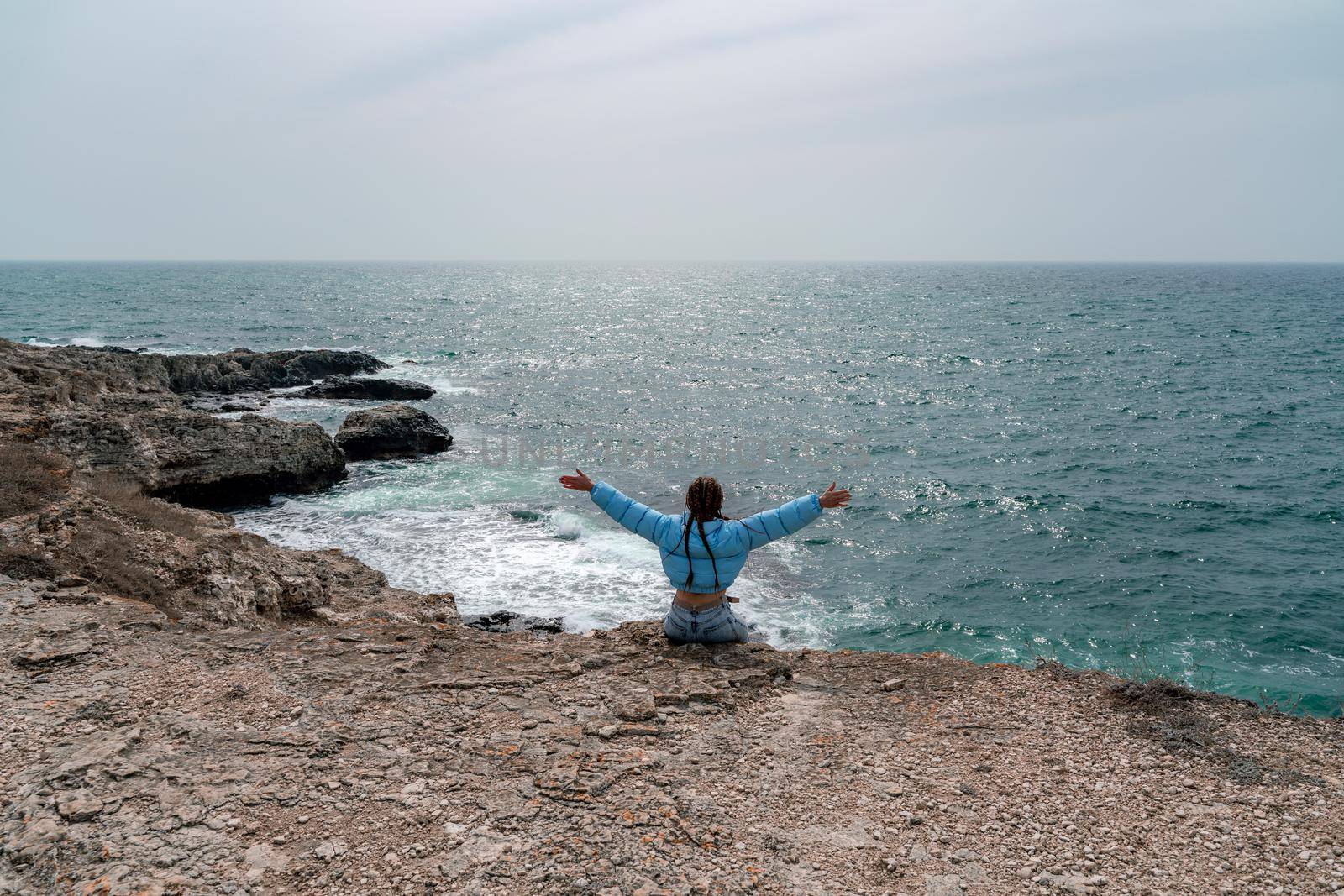 A woman in a blue jacket sits on a rock above a cliff above the sea, looking at the stormy ocean. Girl traveler rests, thinks, dreams, enjoys nature. Peace and calm landscape, windy weather