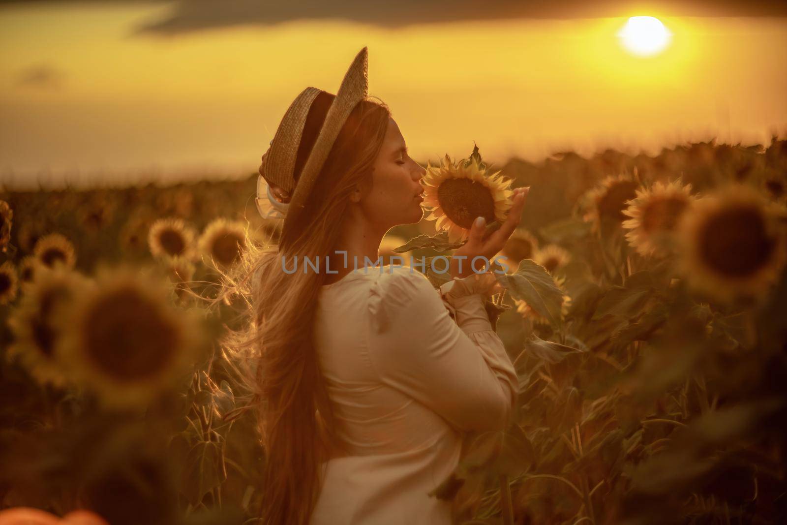 Beautiful middle aged woman looks good in a hat enjoying nature in a field of sunflowers at sunset. Summer. Attractive brunette with long healthy hair. by Matiunina