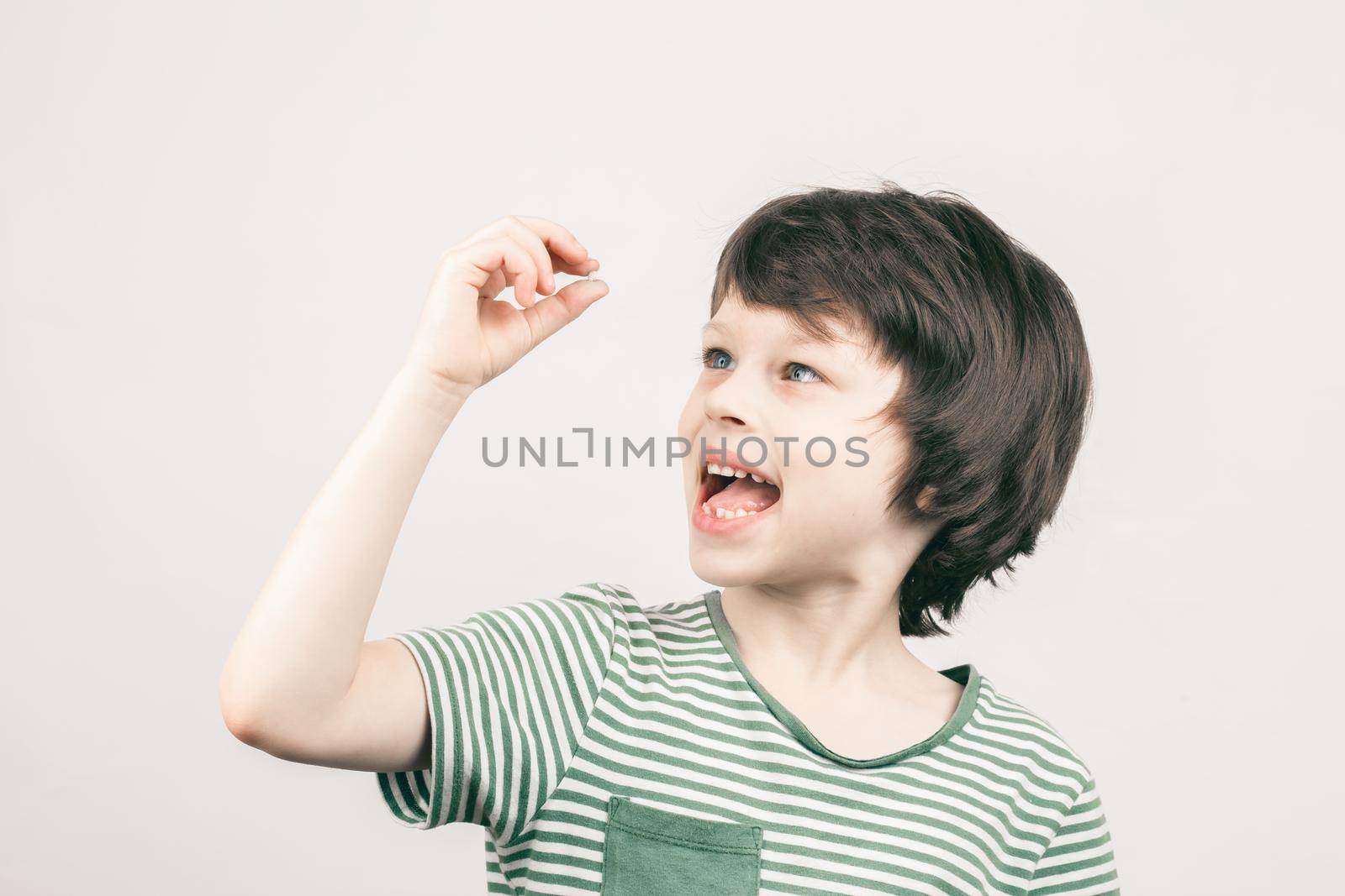 Young toothless boy looking at his first lost bottom front milk tooth over grey background. Child dental health care concept.