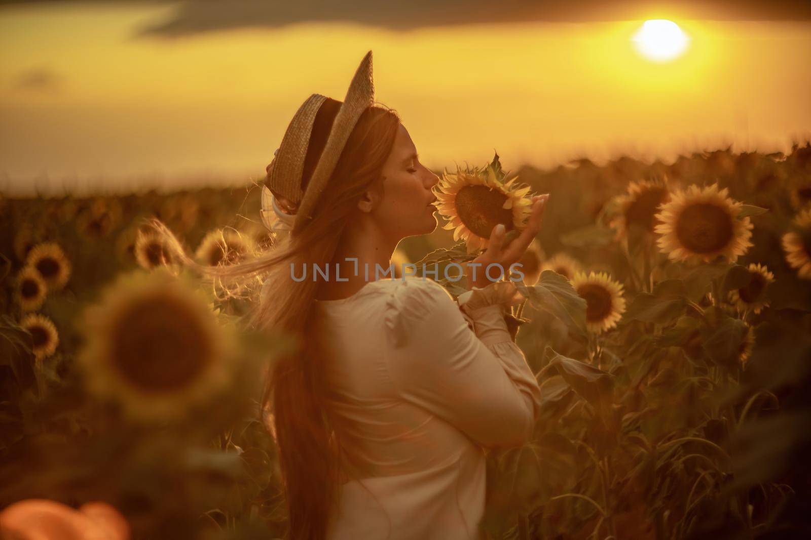 Beautiful middle aged woman looks good in a hat enjoying nature in a field of sunflowers at sunset. Summer. Attractive brunette with long healthy hair. by Matiunina