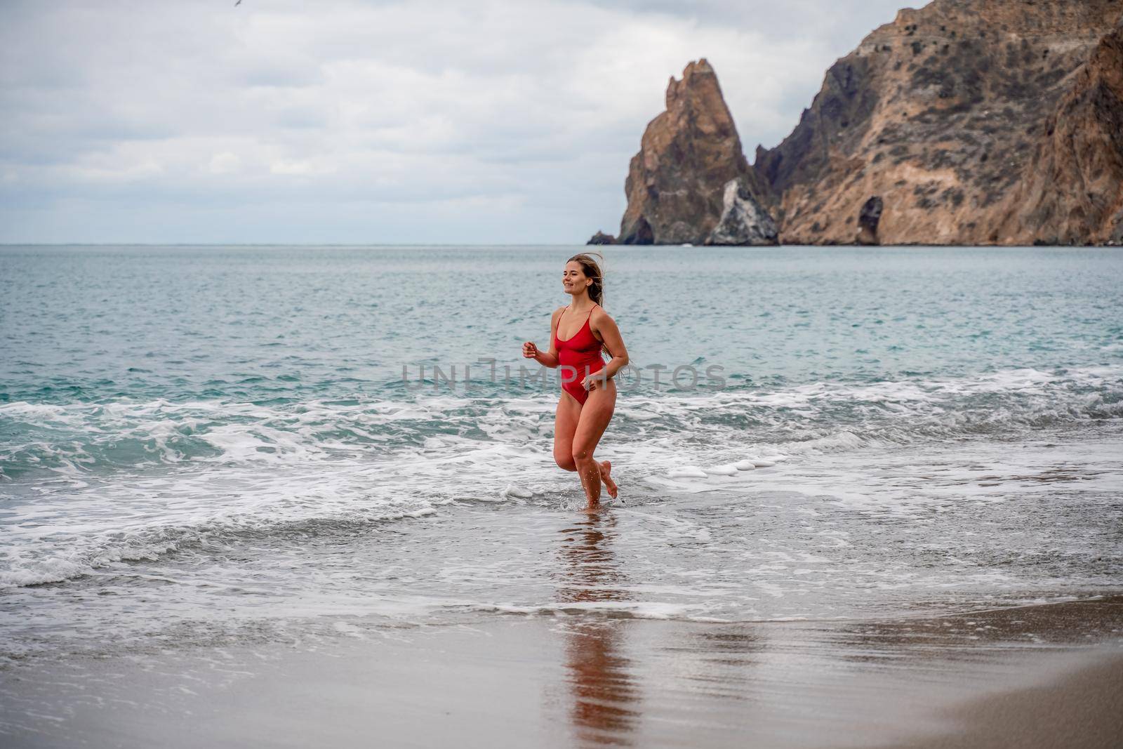 A beautiful and sexy brunette in a red swimsuit on a pebble beach, Running along the shore in the foam of the waves by Matiunina