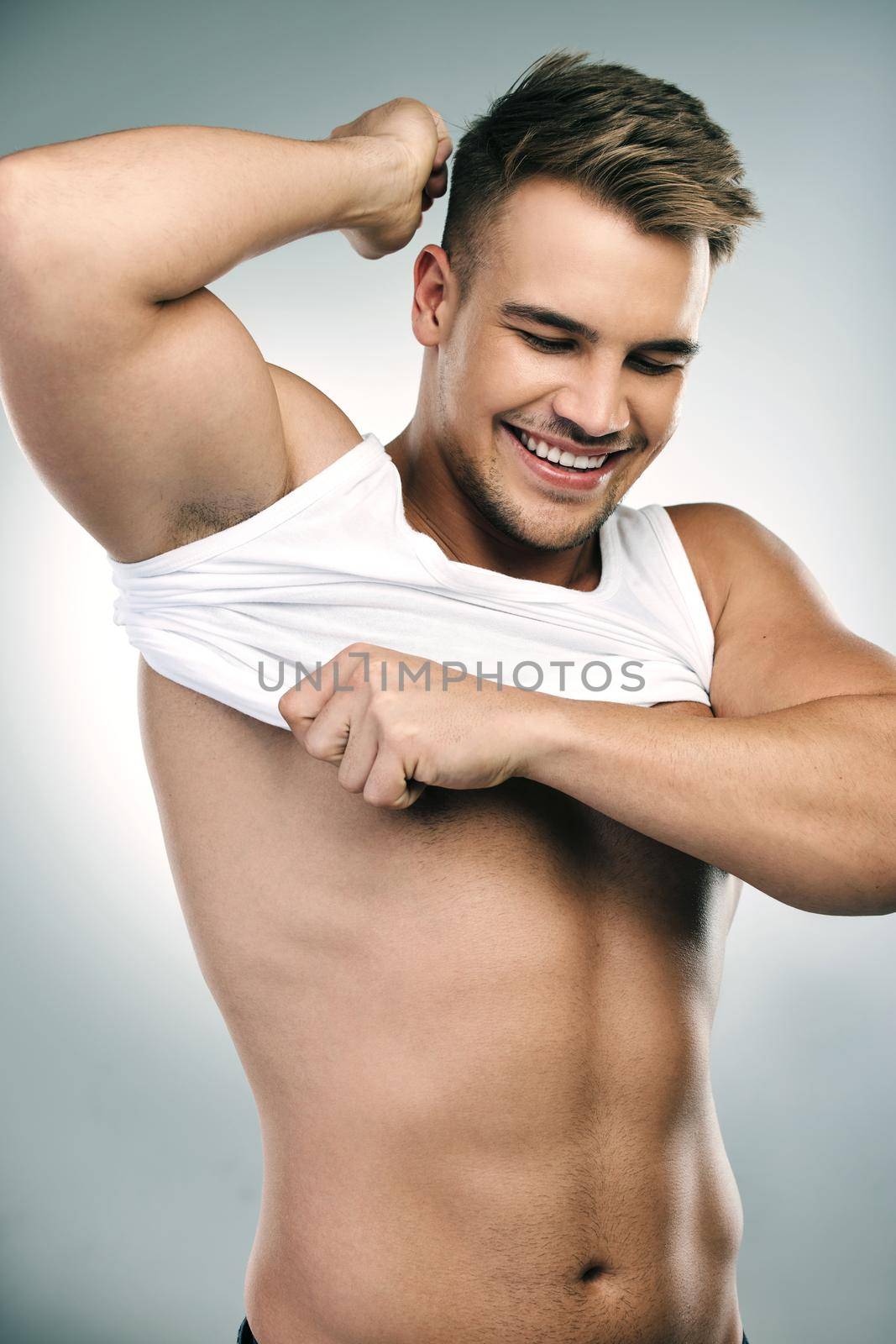 Studio shot of a handsome young man taking his tank top off against a grey background.