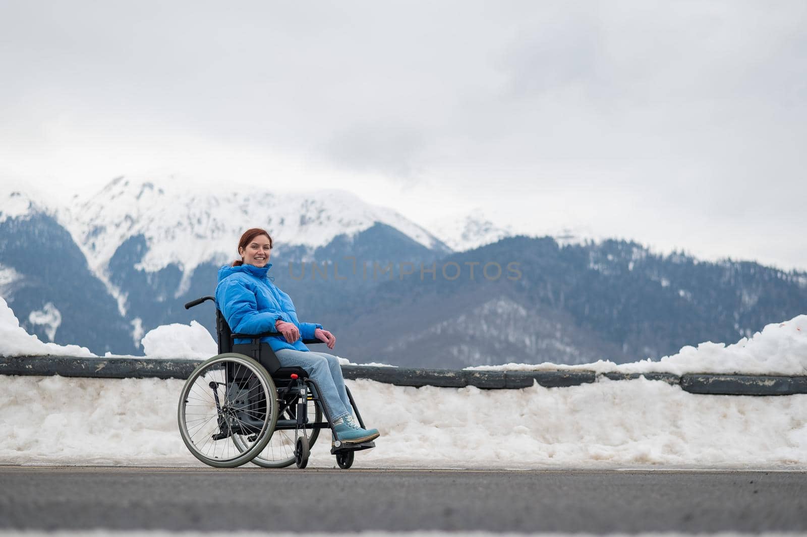 A happy woman dressed in a blue coat sits in a wheelchair on a point view and looking at the snow-capped mountains