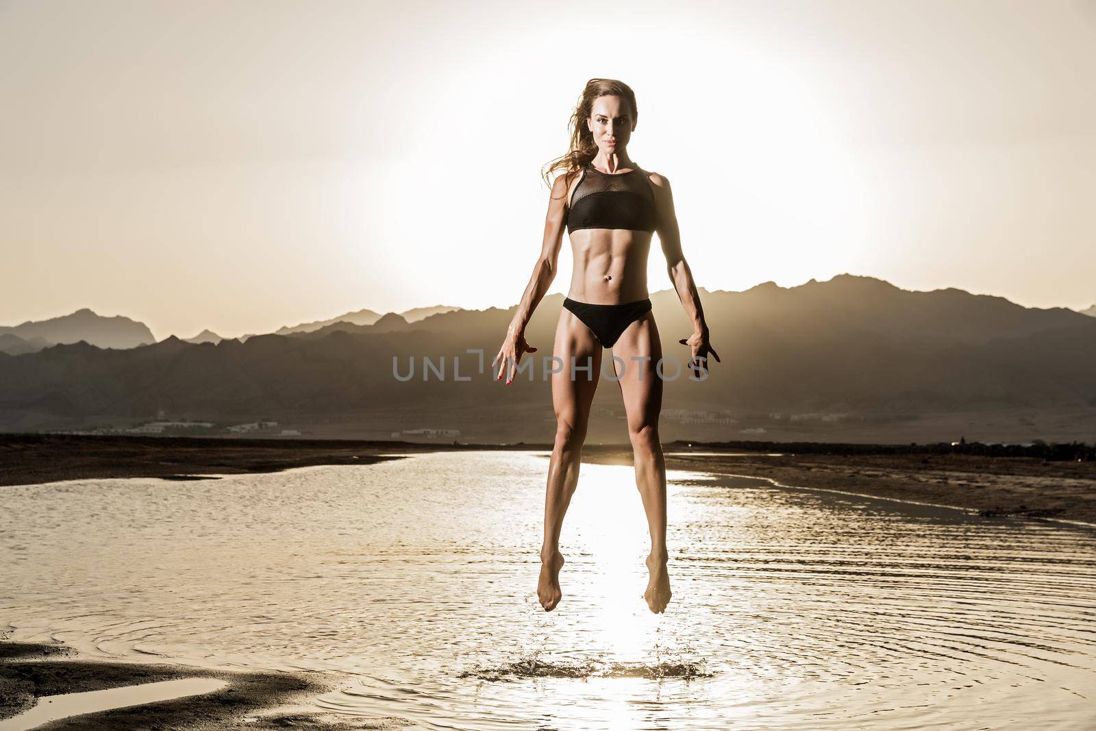 The beautiful girl with a sports body jumps on pool, is dressed in a black swimsuit, long curly hair, pirouettes from track and field athletics, mountain and sandy barkhans are on background. High quality photo