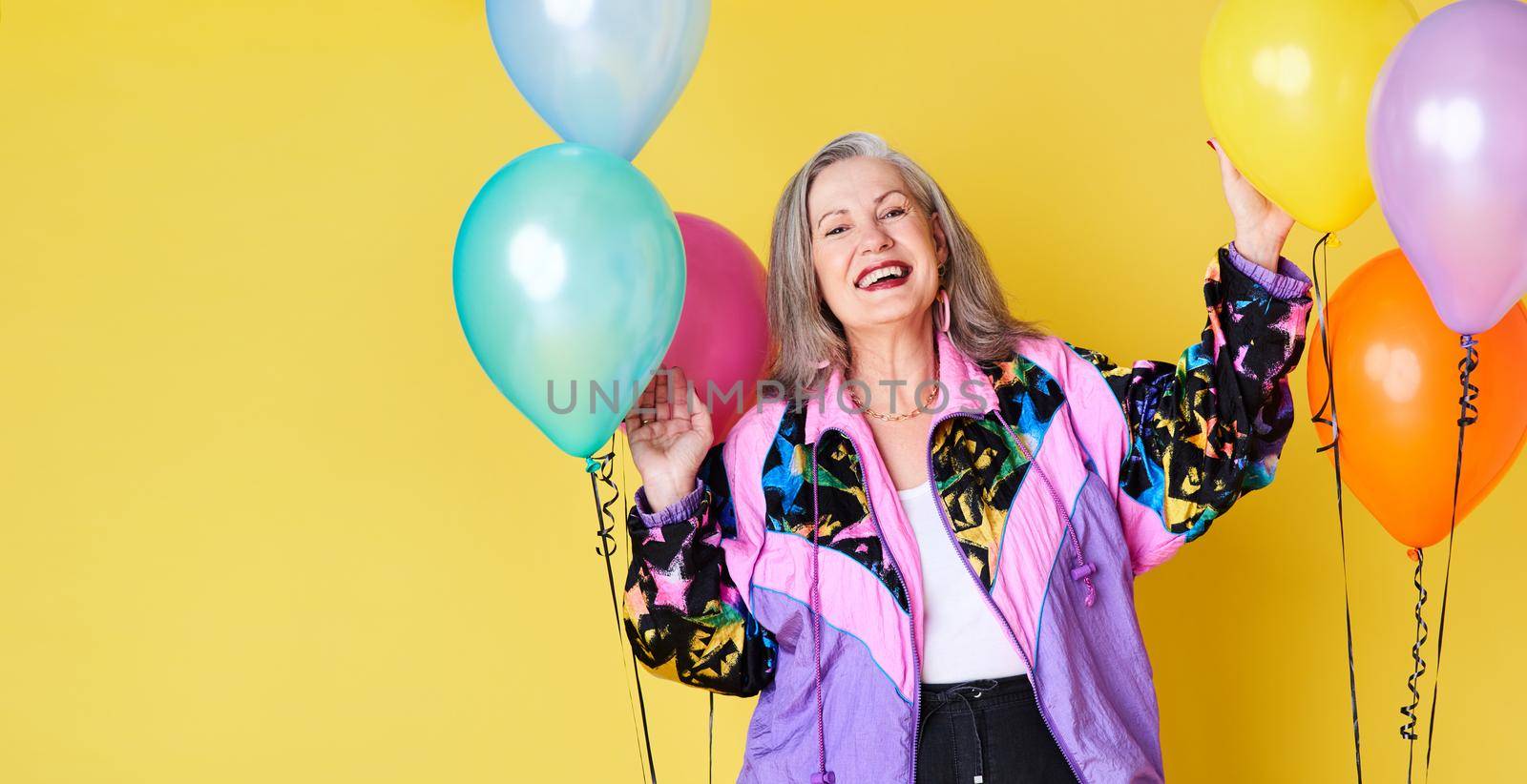 Portrait of a cheerful and stylish senior woman posing with balloons against a yellow background.