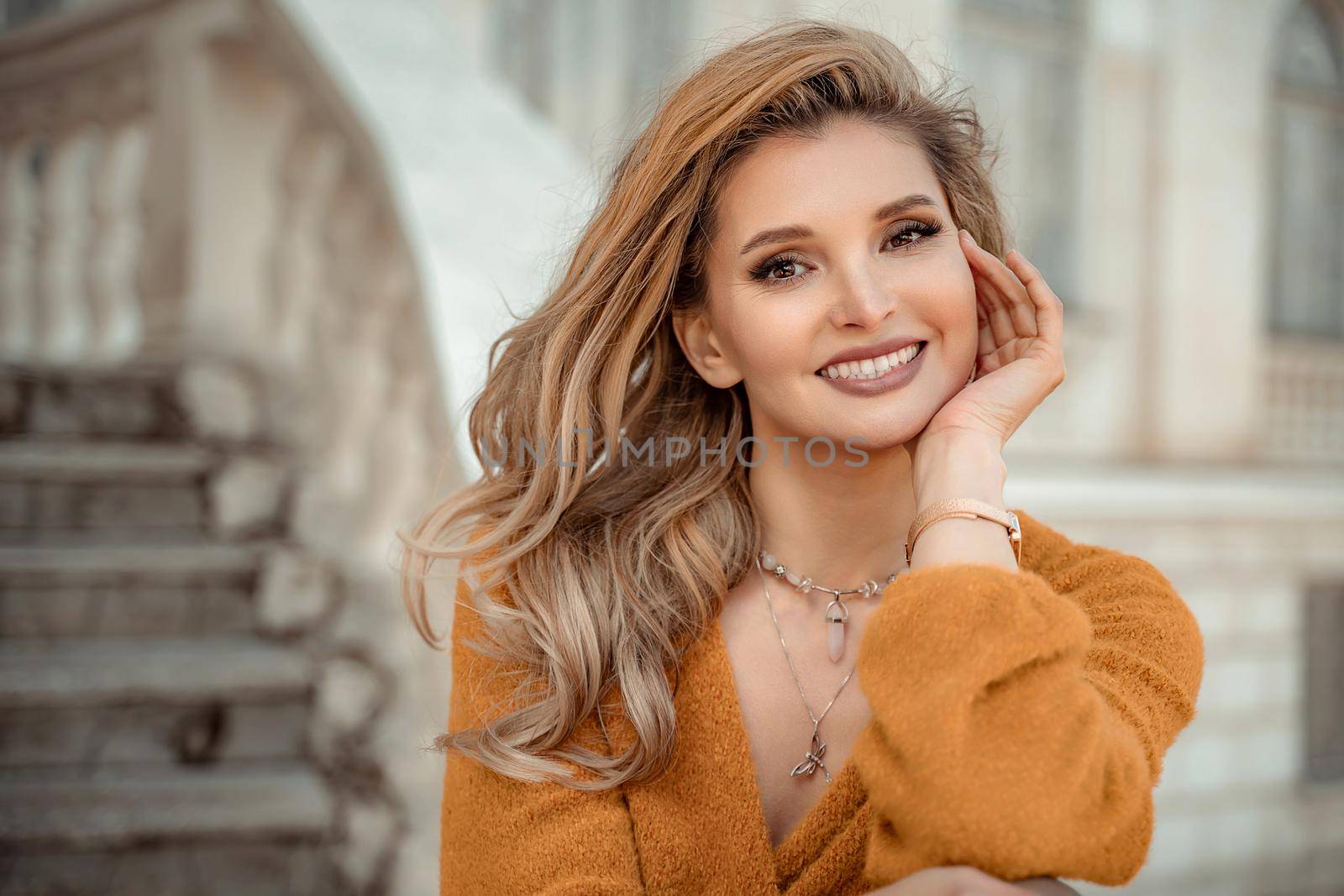 A middle-aged woman looks good blonde with curly beautiful hair and make-up on the background of the building. She smiles openly and looks into the camera. by Matiunina