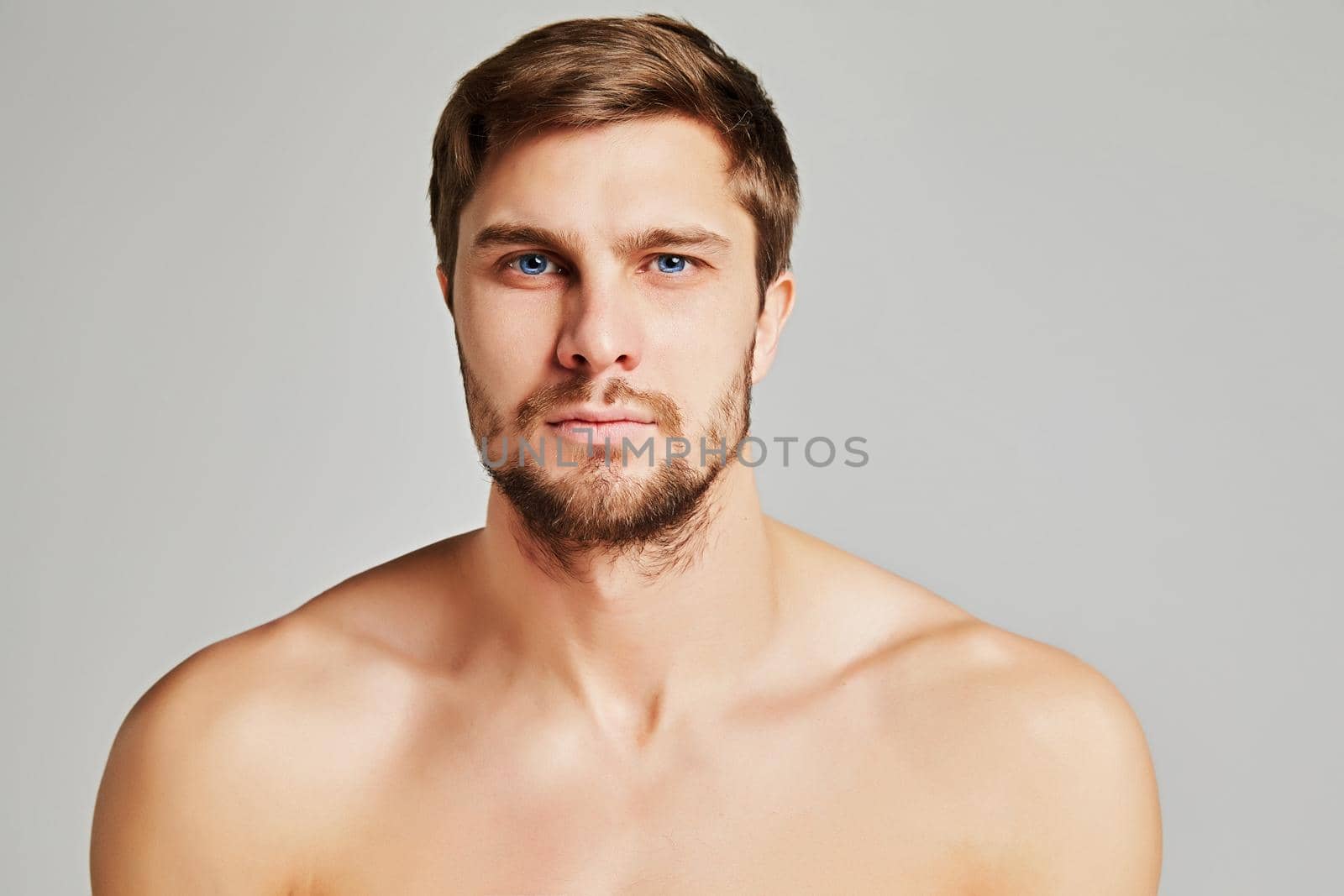 Portrait of a serious young man with bare shoulders on a gray background, powerful swimmers shoulders, beard, charismatic, adult, brutal, athletic. High quality photo